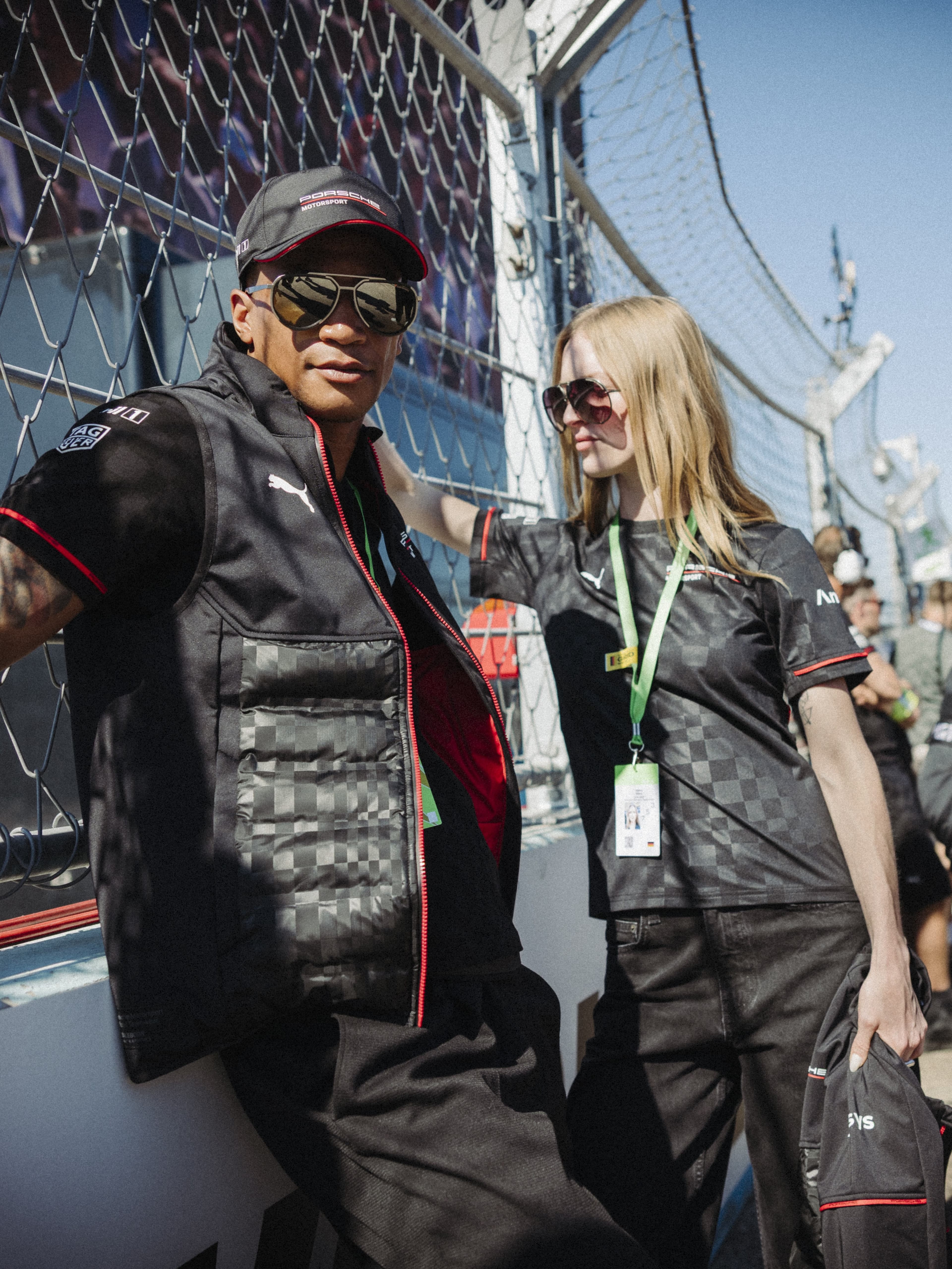 Two people wearing Porsche Motorsport clothing and sunglasses at the racetrack fence under a blue sky.