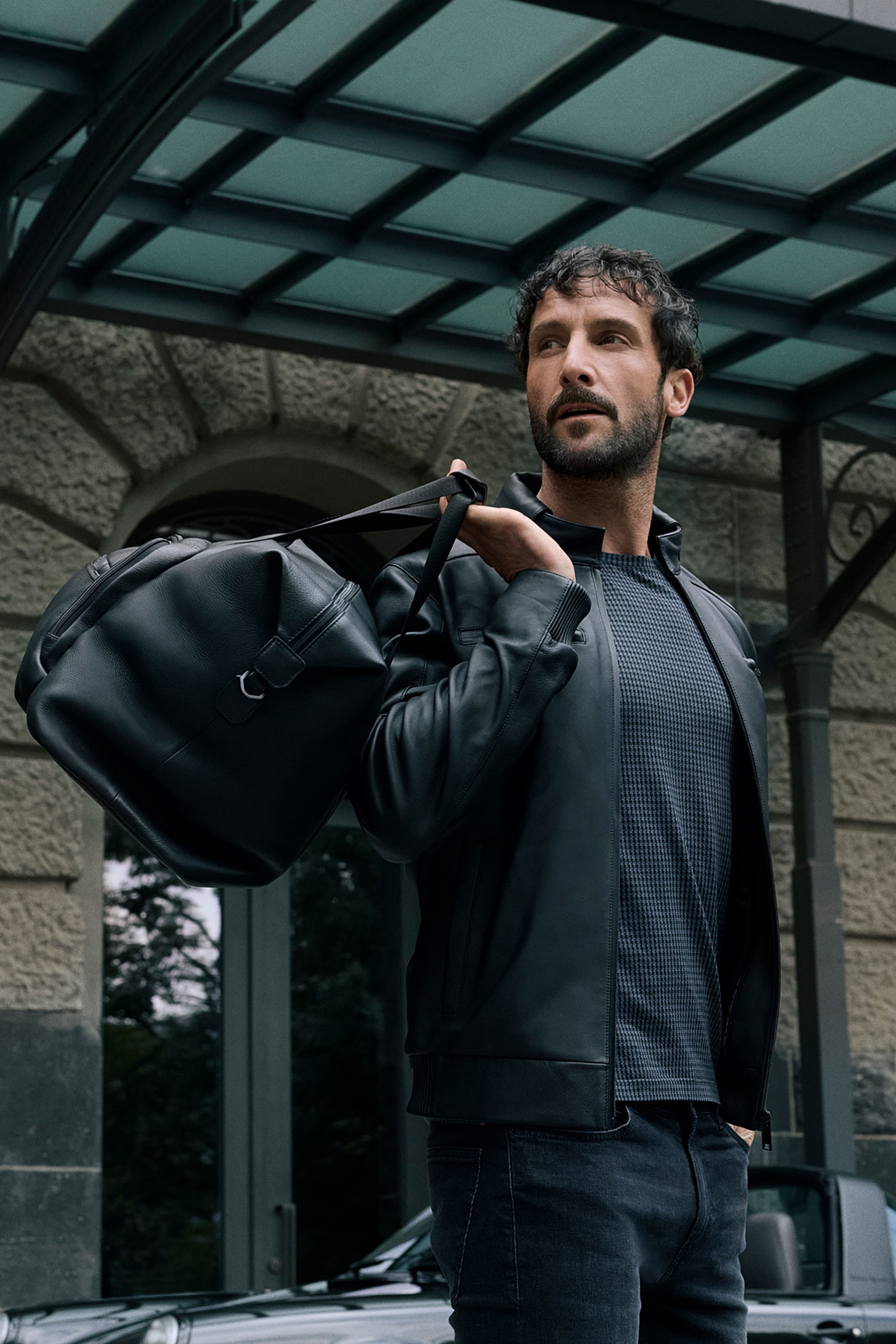 Uomo con giacca in pelle nera che porta una borsa da viaggio nera a tracolla sotto una pensilina in vetro, all’esterno di un edificio