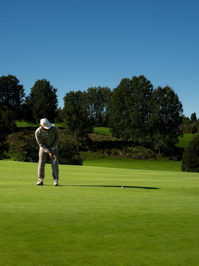 Golfista com roupa bege e boné branco a fazer um putt num green, sob céu azul limpo, com árvores ao fundo.