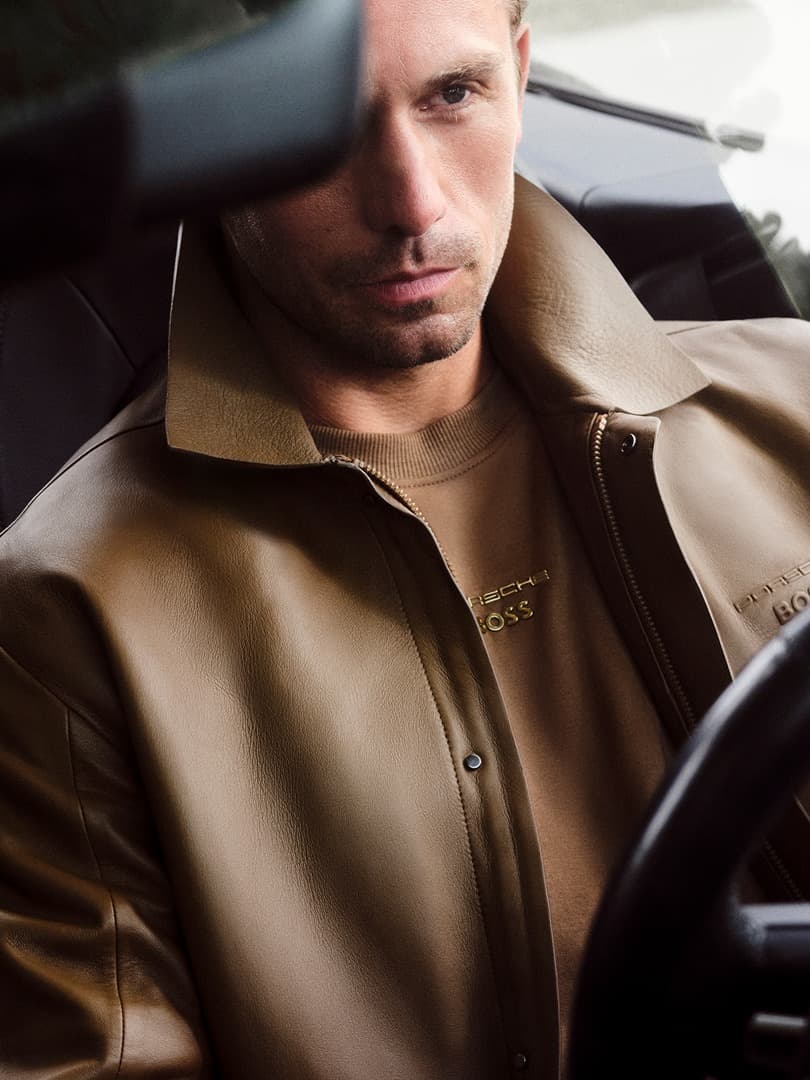 Uomo in auto con giacca in pelle marrone e maglietta beige, primo piano attraverso il parabrezza.