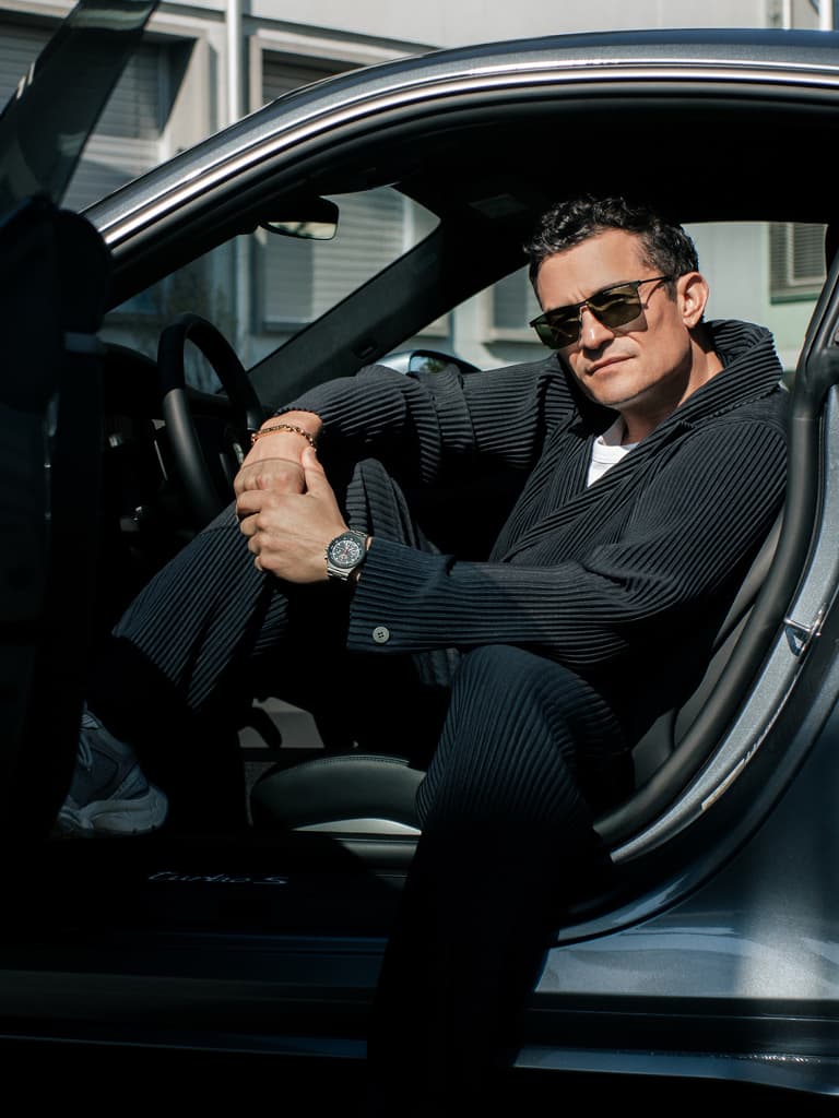 Man wearing sunglasses and a wristwatch sits in the open door of a grey Porsche, wearing a dark suit.
