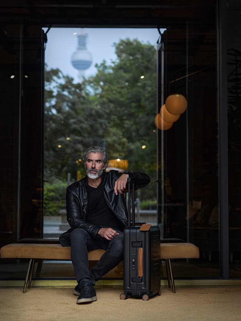 Man in a black leather jacket sitting on a bench, with a black hard-shell suitcase with telescopic handle beside him.
