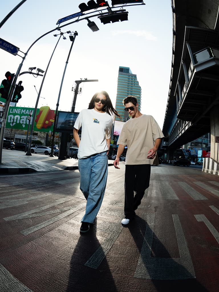 Zwei Personen mit Sonnenbrillen in Oversize-T-Shirts und Jeans/Schwarz, gehen über einen Zebrastreifen in der Stadt.