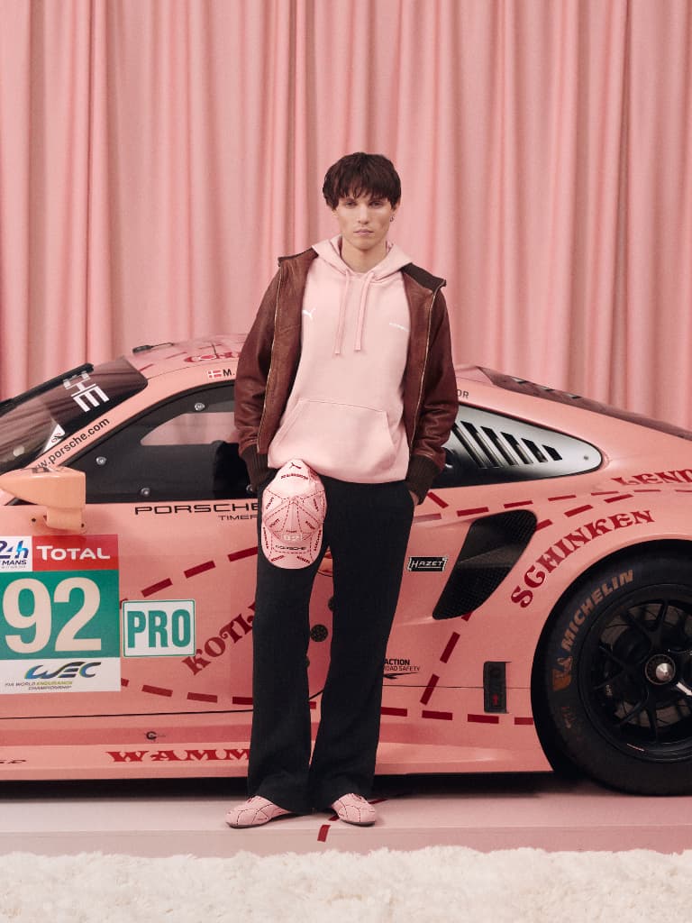 Person in pink hoodie and brown jacket stands by a pink Porsche race car with matching cap and shoes.