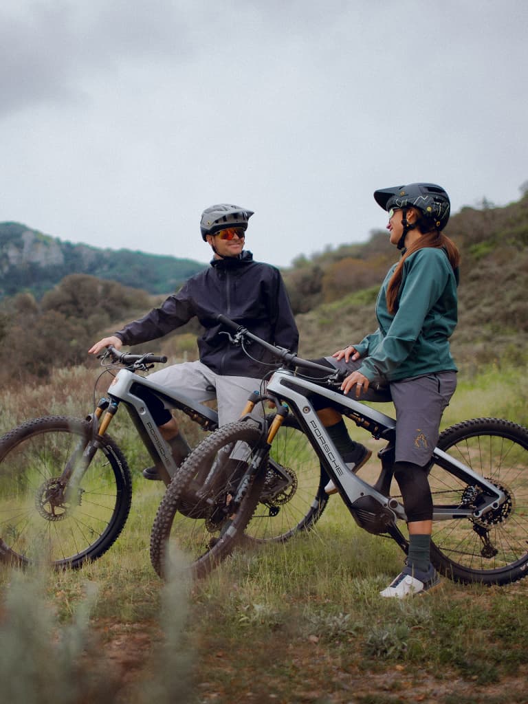 Dois ciclistas com capacetes e equipamento em e-bikes Porsche, a conversar num trilho relvado com colinas ao fundo.