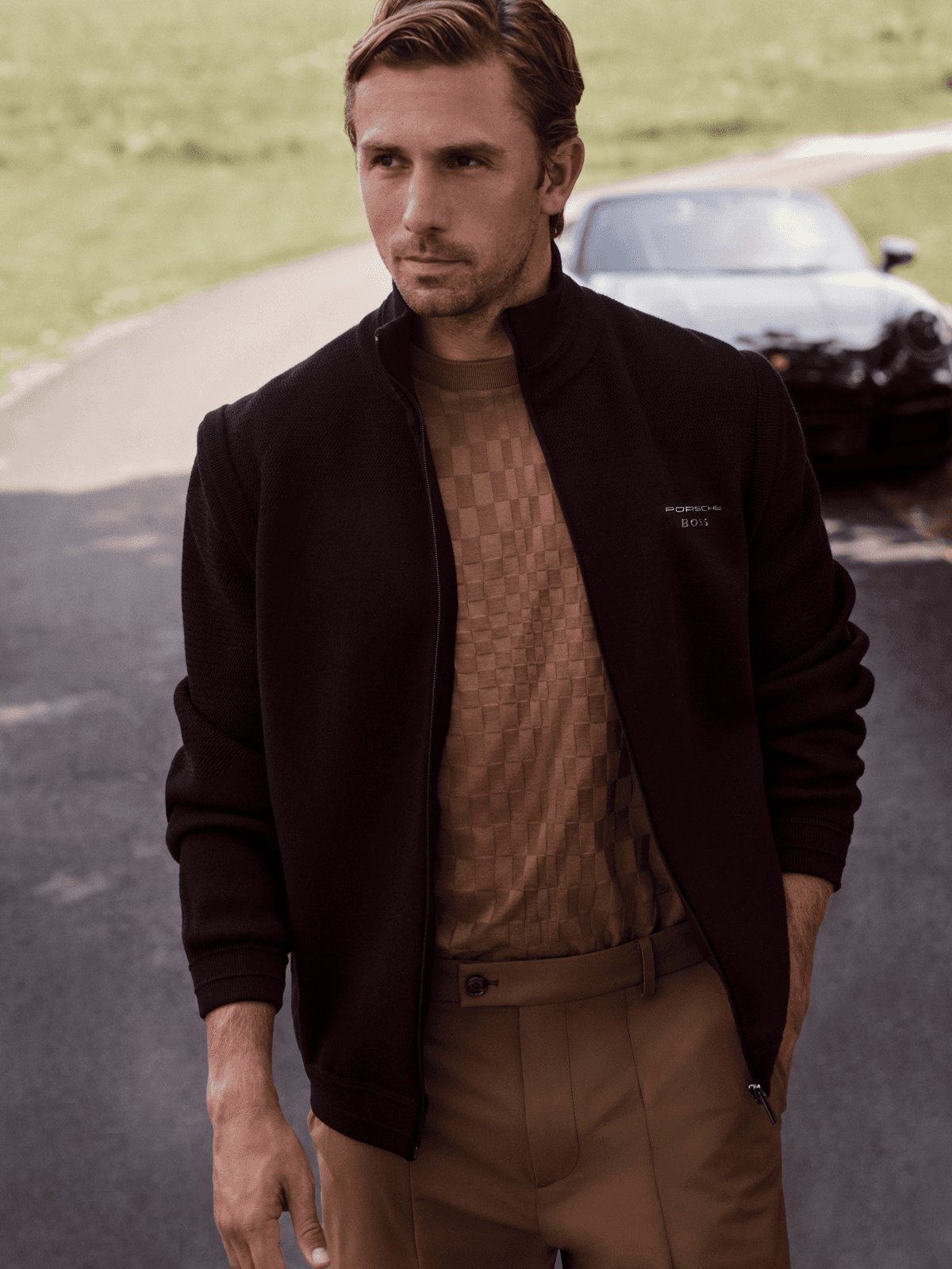 Man wearing a black Porsche BOSS jacket and brown shirt, standing on a country road; Porsche sports car blurred in the background.
