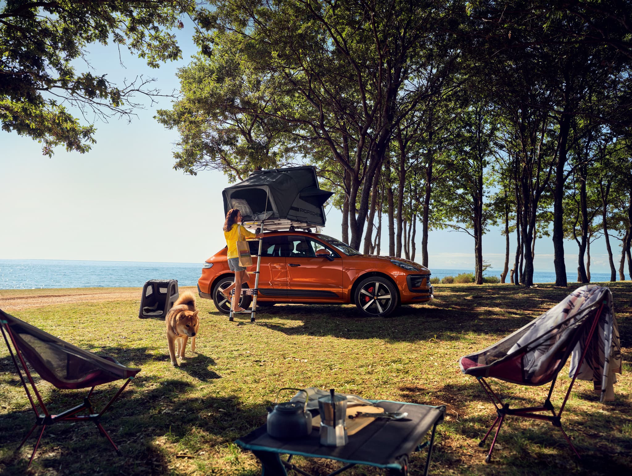 orange Porsche Macan on the grass near to the sea, with a roof tent and some camping equipment