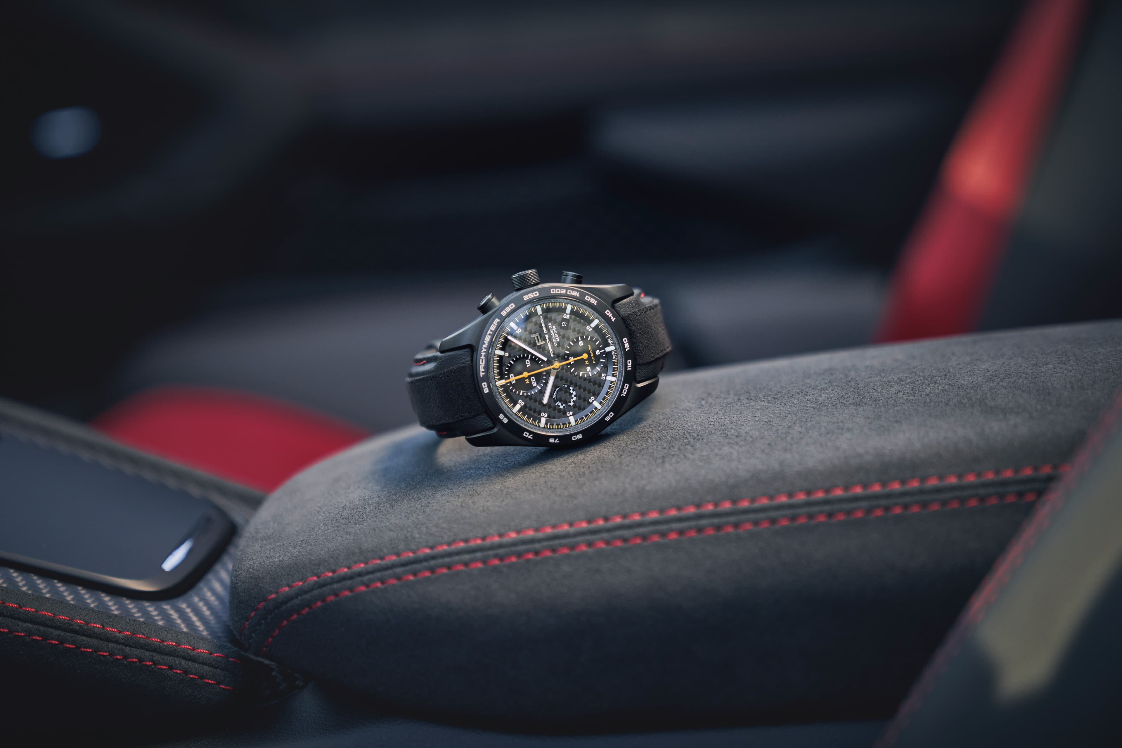 The Porsche Chronograph 718 Spyder RS is shown from the front on a suede seat in a Porsche car
