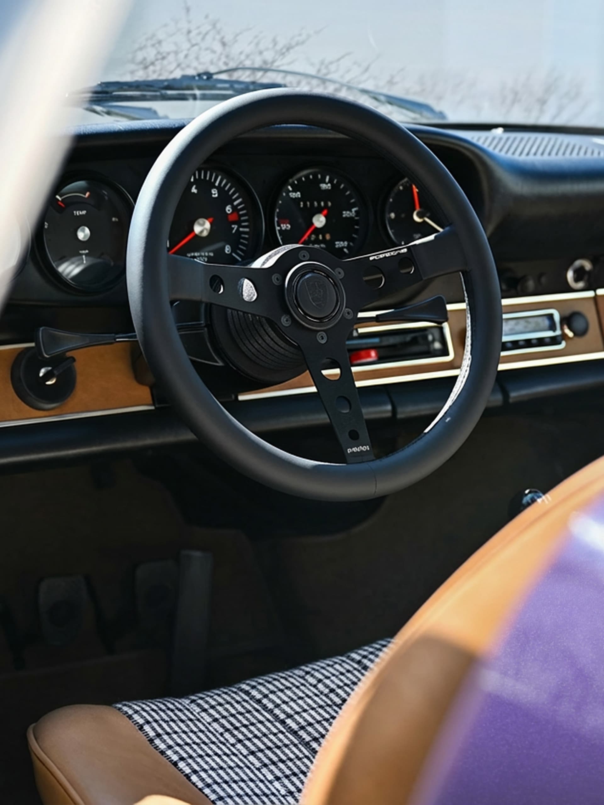 Klassieke auto-interieur met zwart stuurwiel, beige dashboard en geruite zetel. Meters zichtbaar achter het stuur.