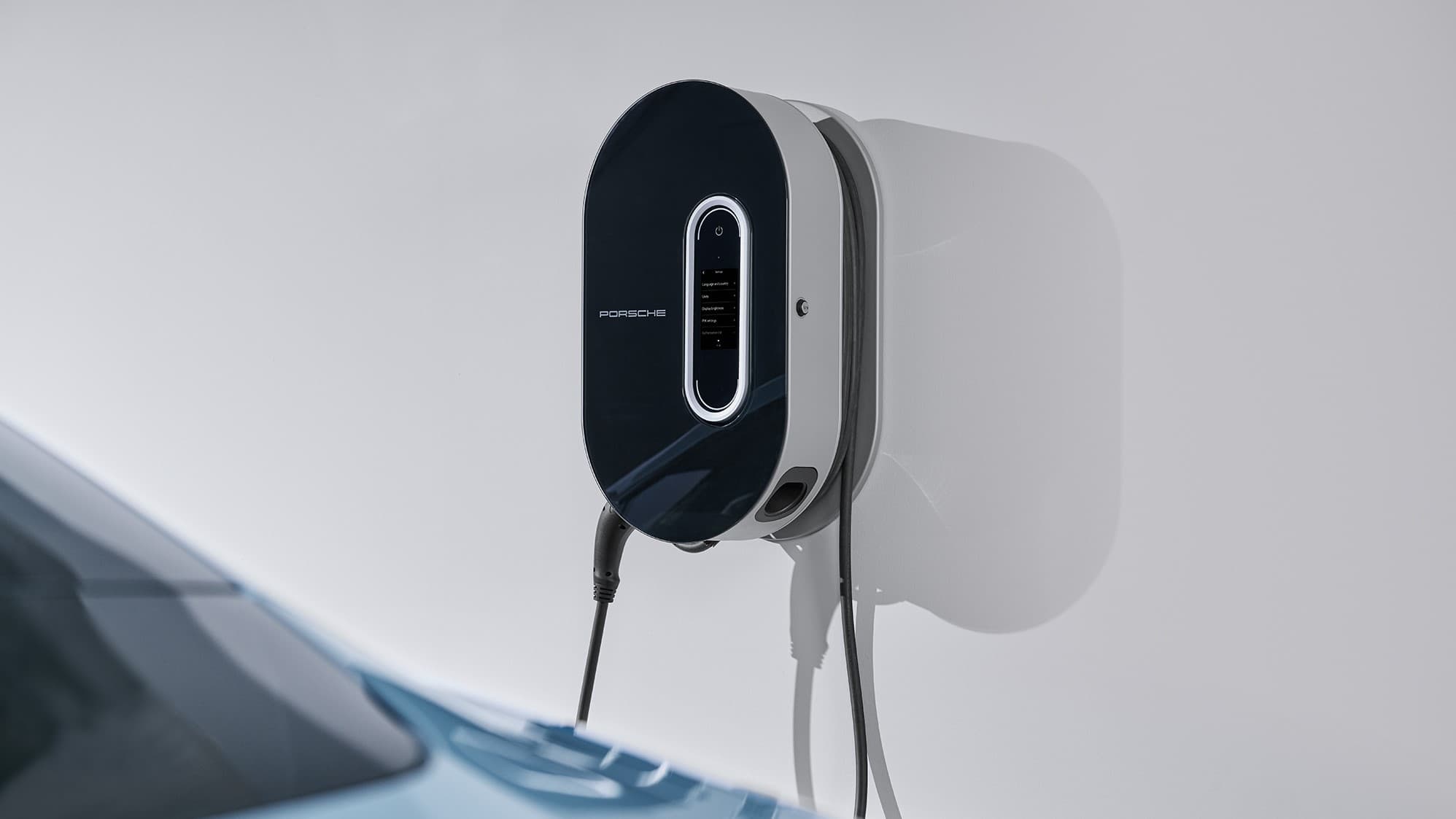 Porsche Wandladegerät in Schwarz-Weiß, montiert an einer weißen Wand, mit einem blauen Auto teilweise im Vordergrund sichtbar.