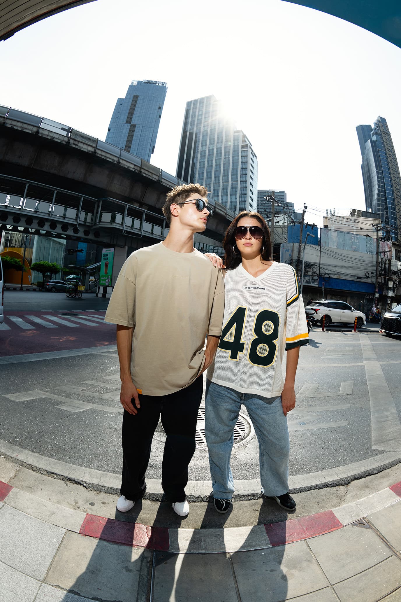 Dos modelos con gafas de sol llevan prendas de la colección Porsche AHEAD: camiseta beige y camiseta tipo jersey blanca de Porsche con el número 48, con rascacielos altos de fondo.