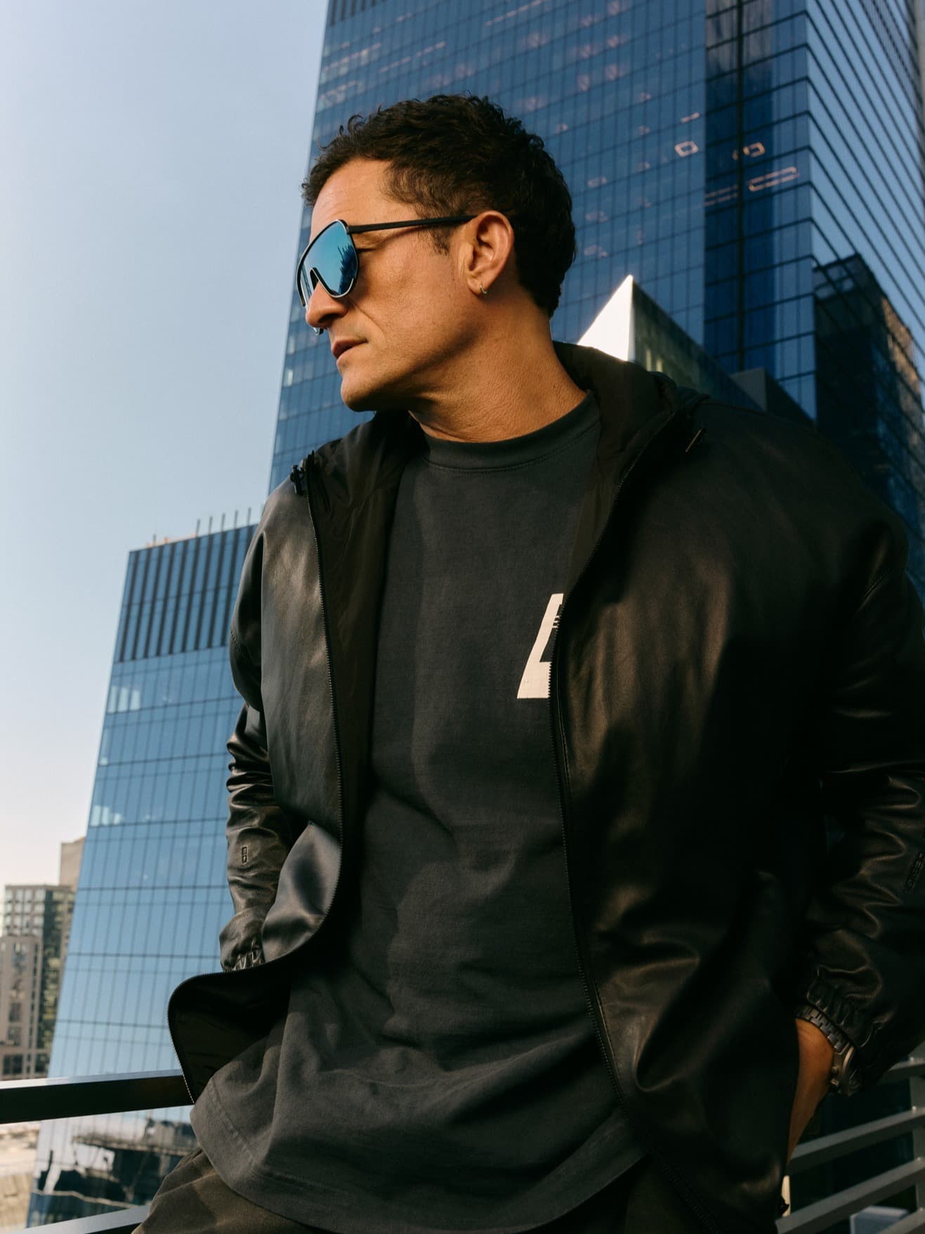 Man wearing mirrored sunglasses and a black leather jacket in front of glass high-rise buildings in the city.