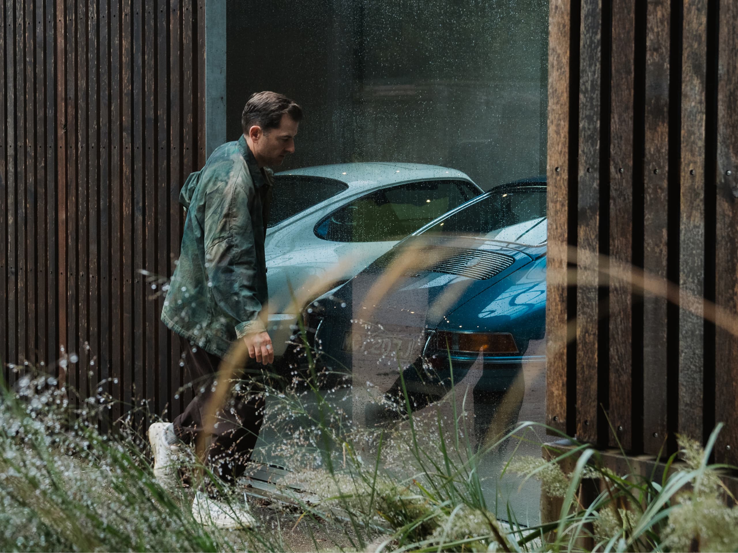 Man in green jacket walking past a window with two classic cars, one white and one blue, visible through rain-speckled glass.
