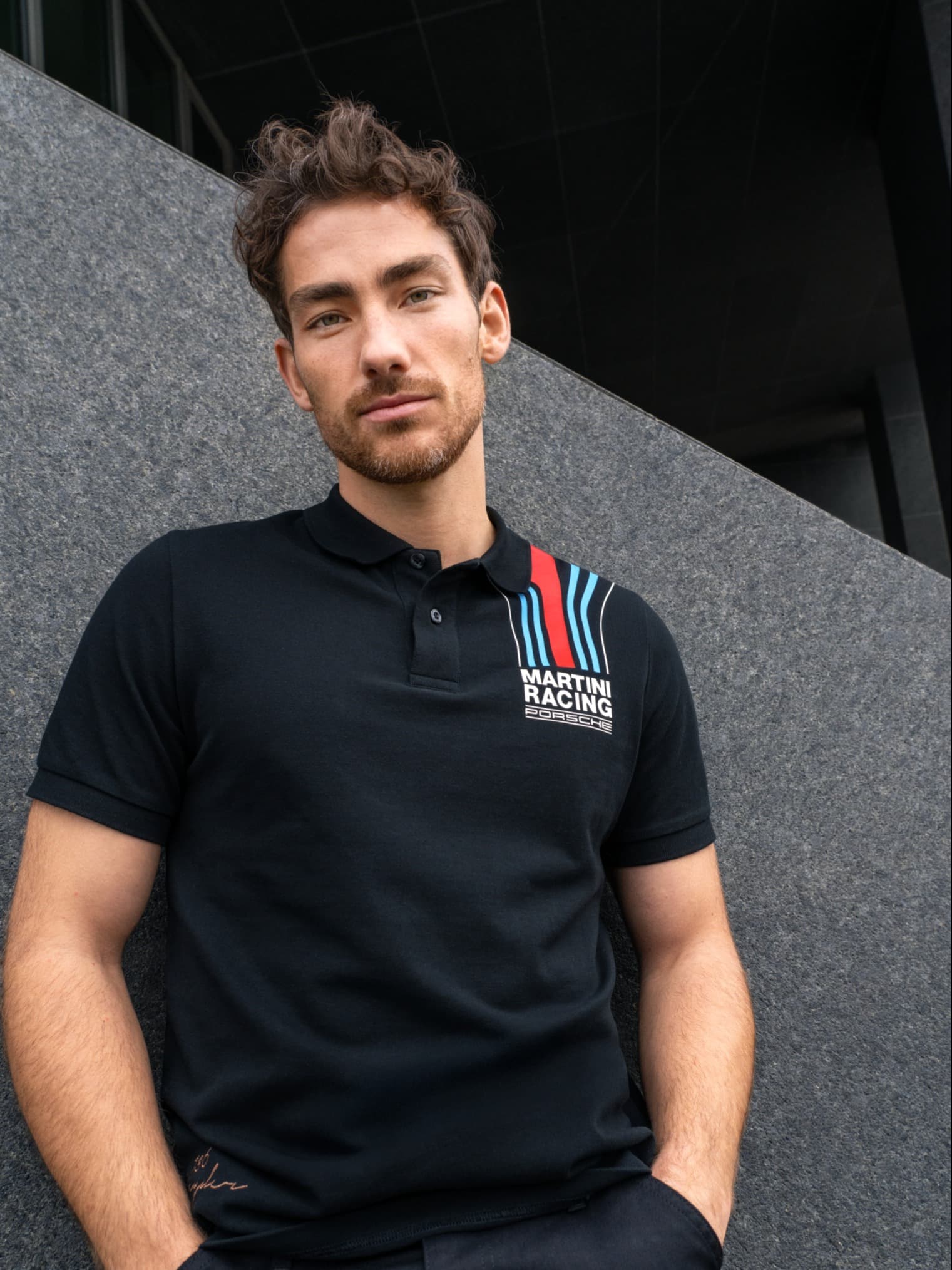 Mann im schwarzen Porsche Martini Racing Polohemd, steht vor einer grauen Wand und schaut in die Kamera.