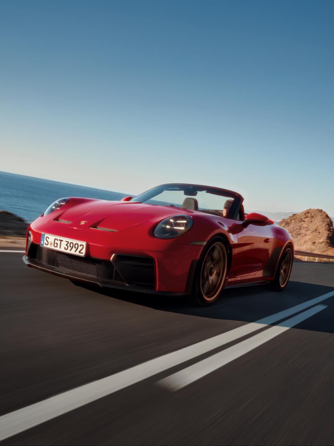 Rød Porsche 911 Cabriolet kører på kystvej, hav til venstre og klipper til højre under blå himmel.
