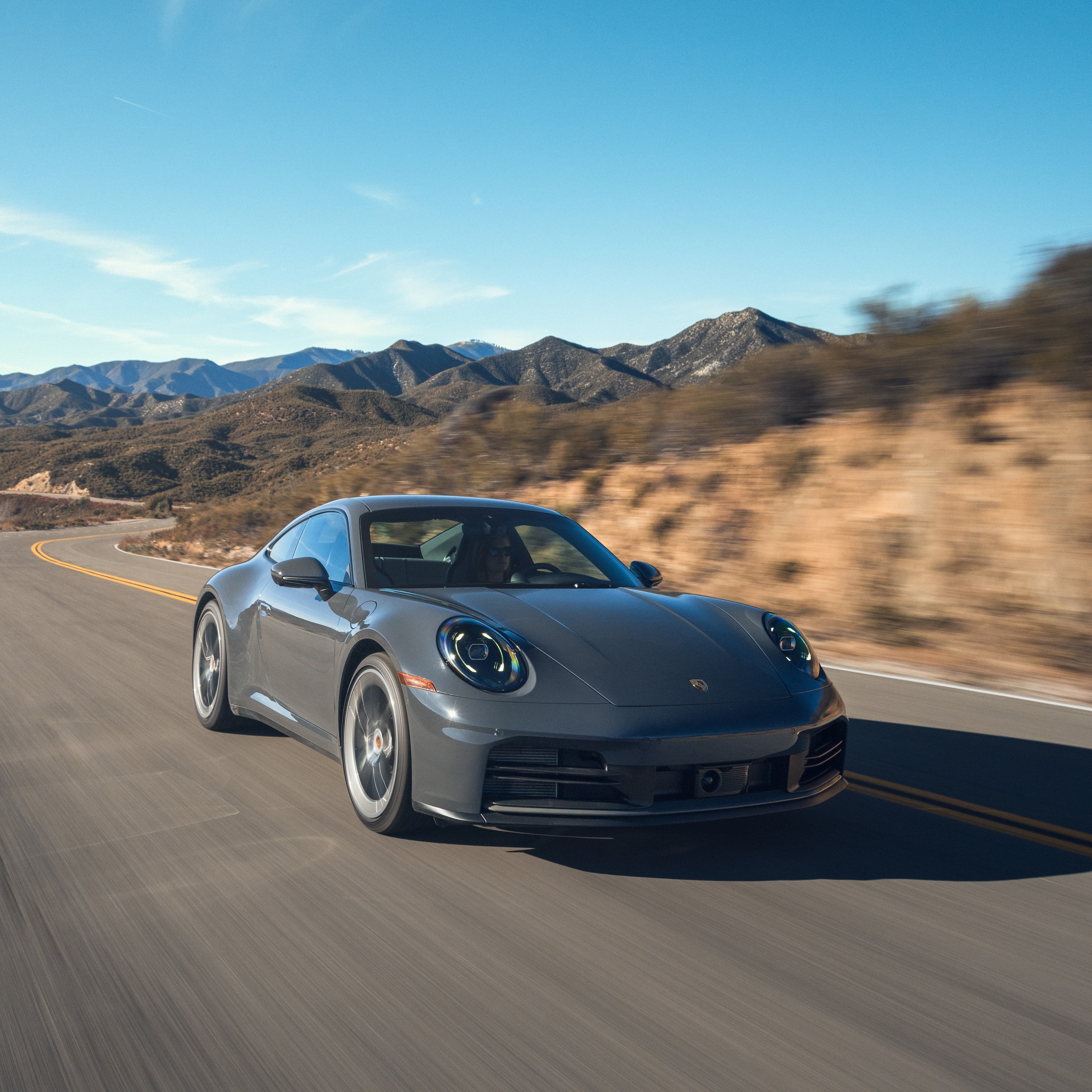 Gray Porsche 911 driving on a winding country road, with mountains and blue sky in the background.