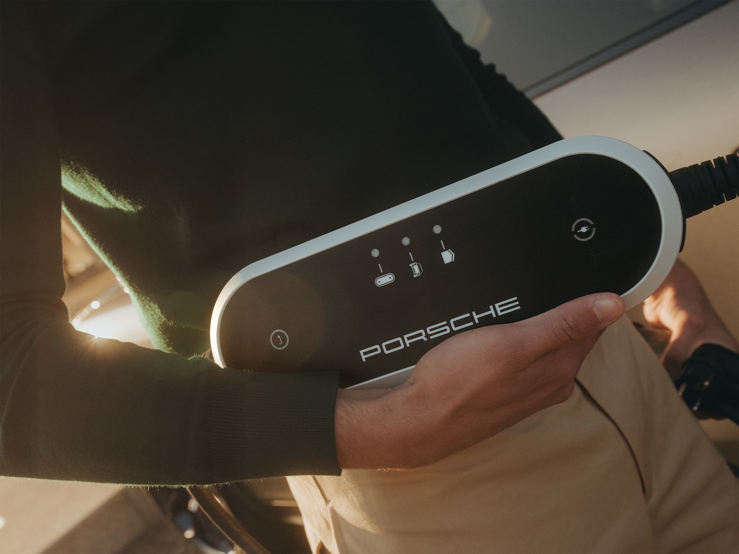 Persona che tiene un dispositivo di ricarica Porsche contro un'auto, indossando un maglione verde scuro e pantaloni beige, alla luce del sole.