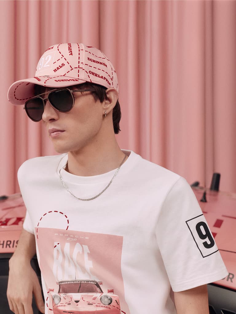Man met roze pet en zonnebril, draagt een wit Porsche t-shirt met een autografiek, tegen een roze achtergrond.