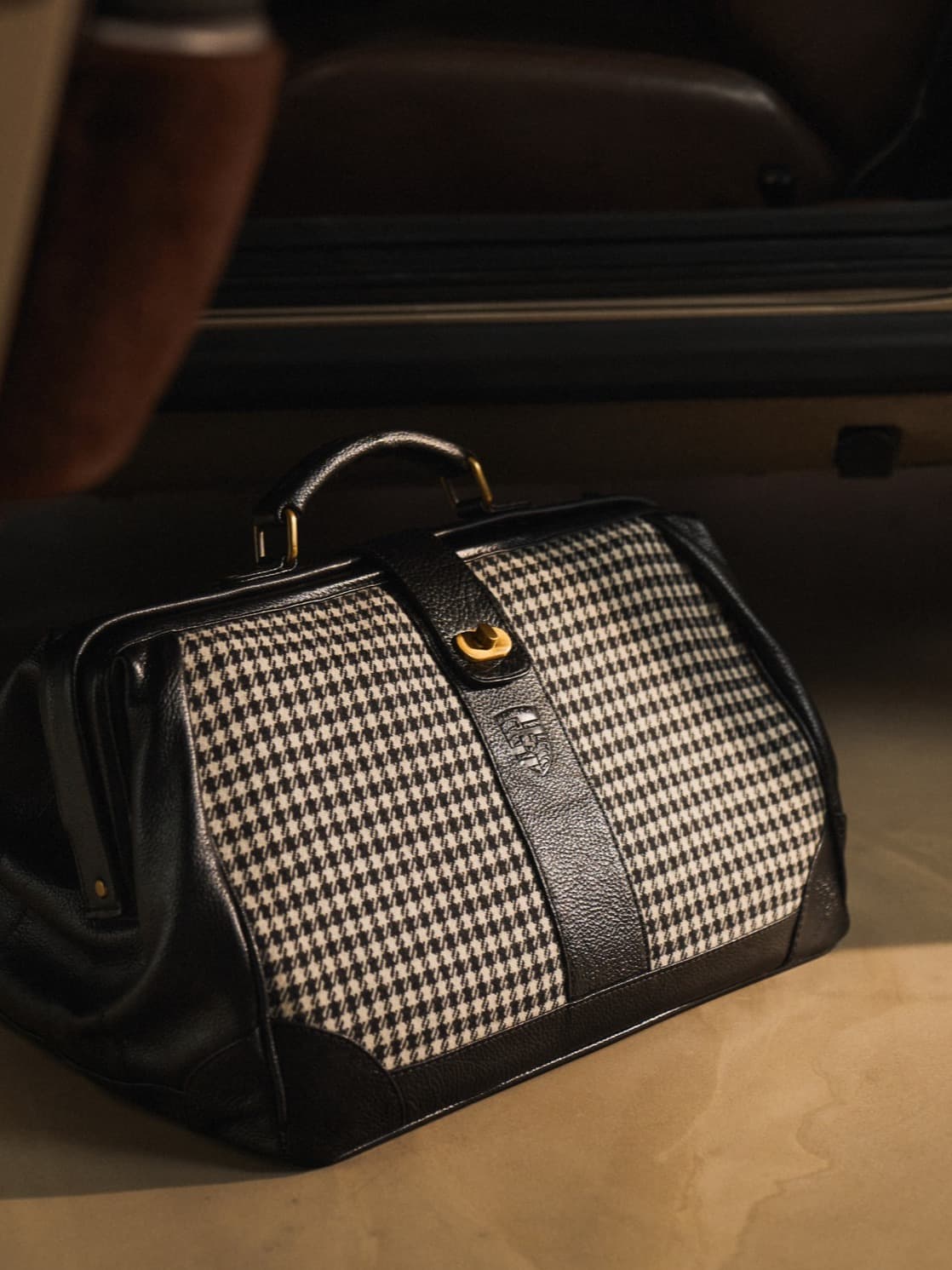 Black-and-white houndstooth travel bag with black leather details and gold hardware, in front of a car door.