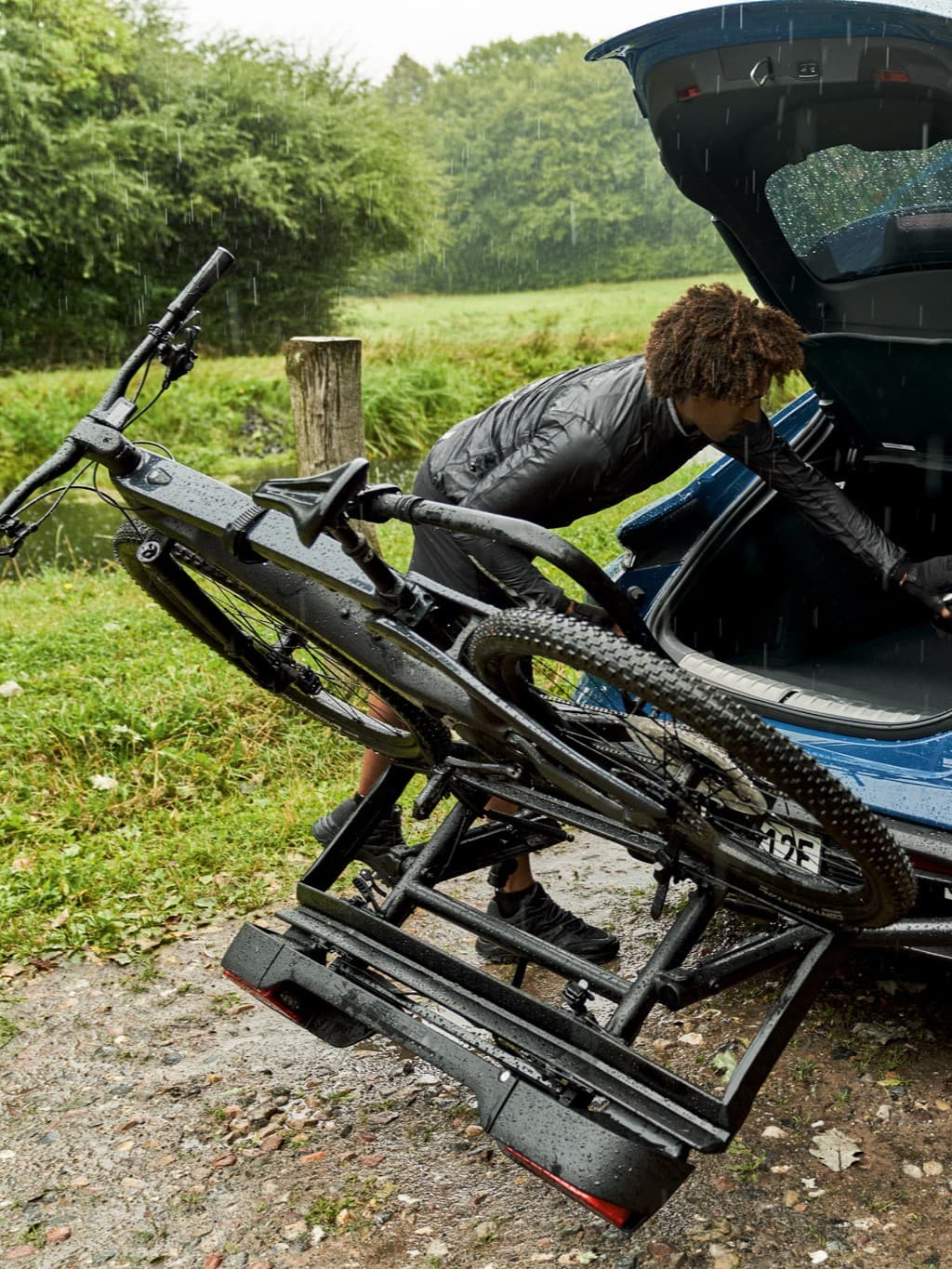 Person lädt ein schwarzes Fahrrad auf einen Auto-Fahrradträger im Regen, umgeben von Grün.