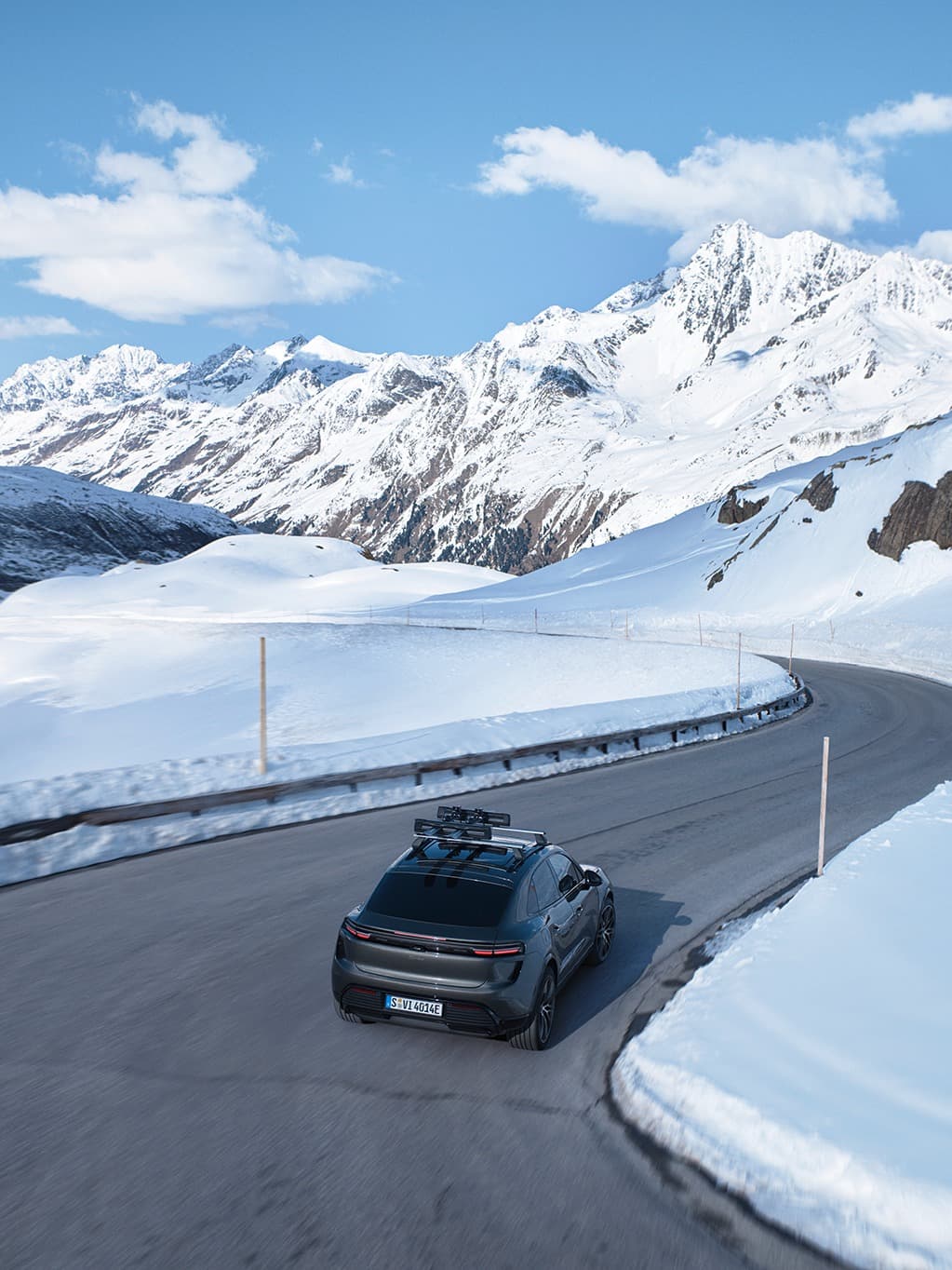 A sleek car drives on a snowy mountain road under a clear blue sky, surrounded by snow-covered peaks.