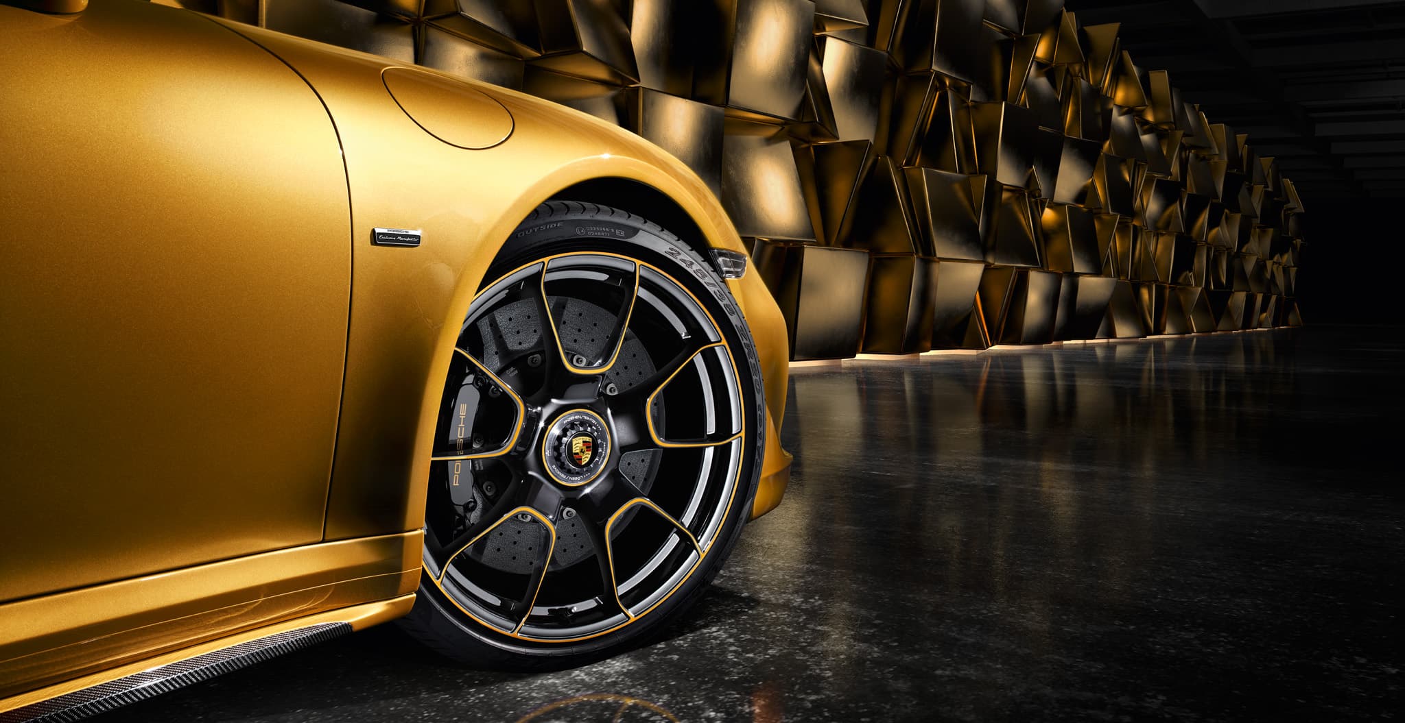 Nahaufnahme des goldenen Porsche-Frontkotflügels und eines schwarzen Leichtmetallrads mit goldenen Akzenten vor einer dunklen geometrischen Wand