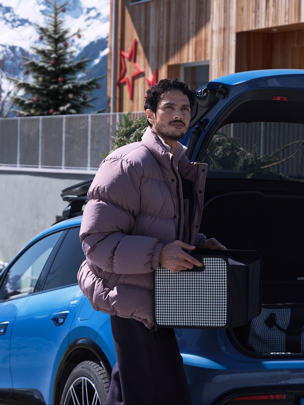 Man in a purple jacket holding a box by a blue car, with a snowy mountain and decorated tree in the background.