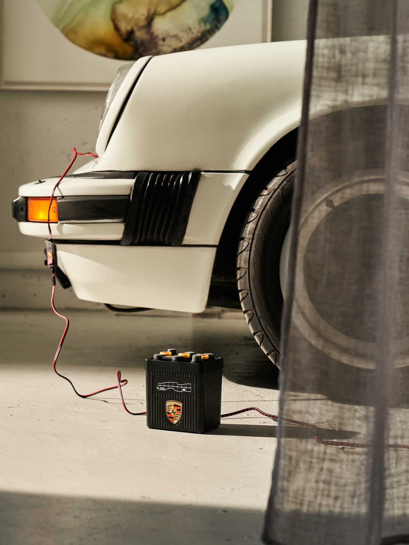 Vit Porsche-bil med ansluten batteriladdare, placerad i ett garage med mjuk belysning och delvis synligt draperi.