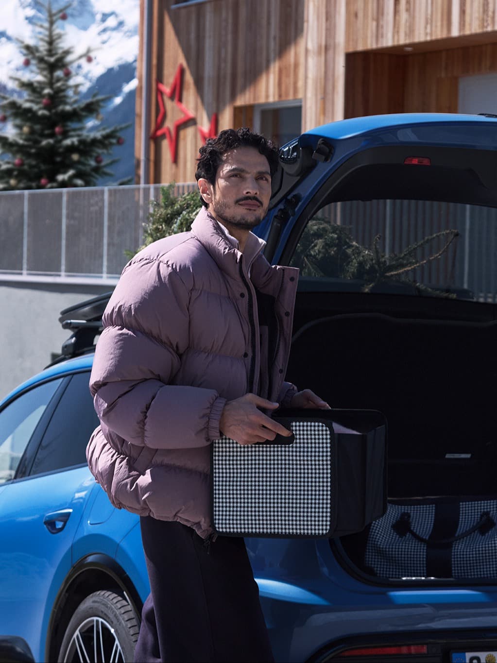 Man in a purple jacket holding a box by a blue car, with a snowy mountain and decorated tree in the background.