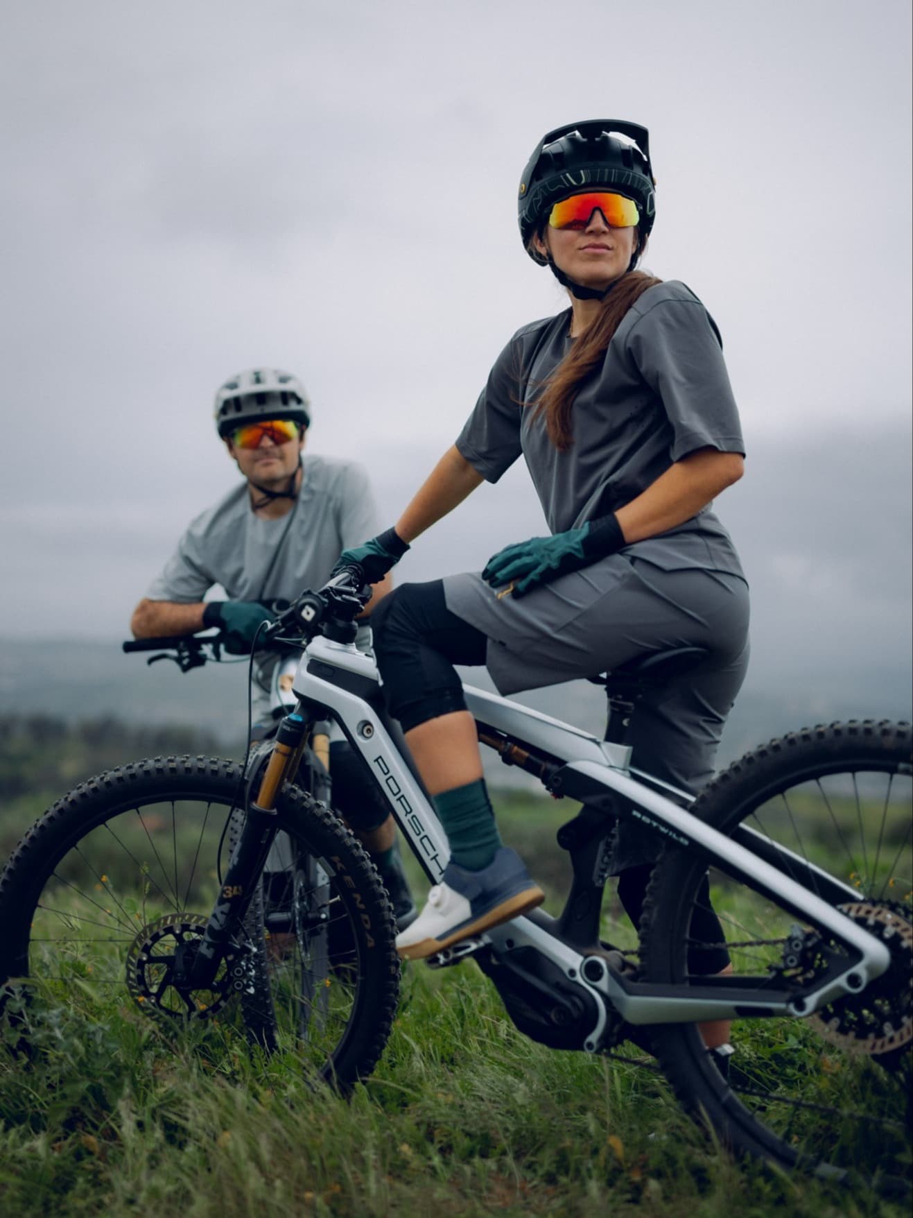 Mountain biker in completo grigio con casco nero e occhiali a specchio su una e-bike Porsche; secondo rider dietro, cielo nuvoloso.
