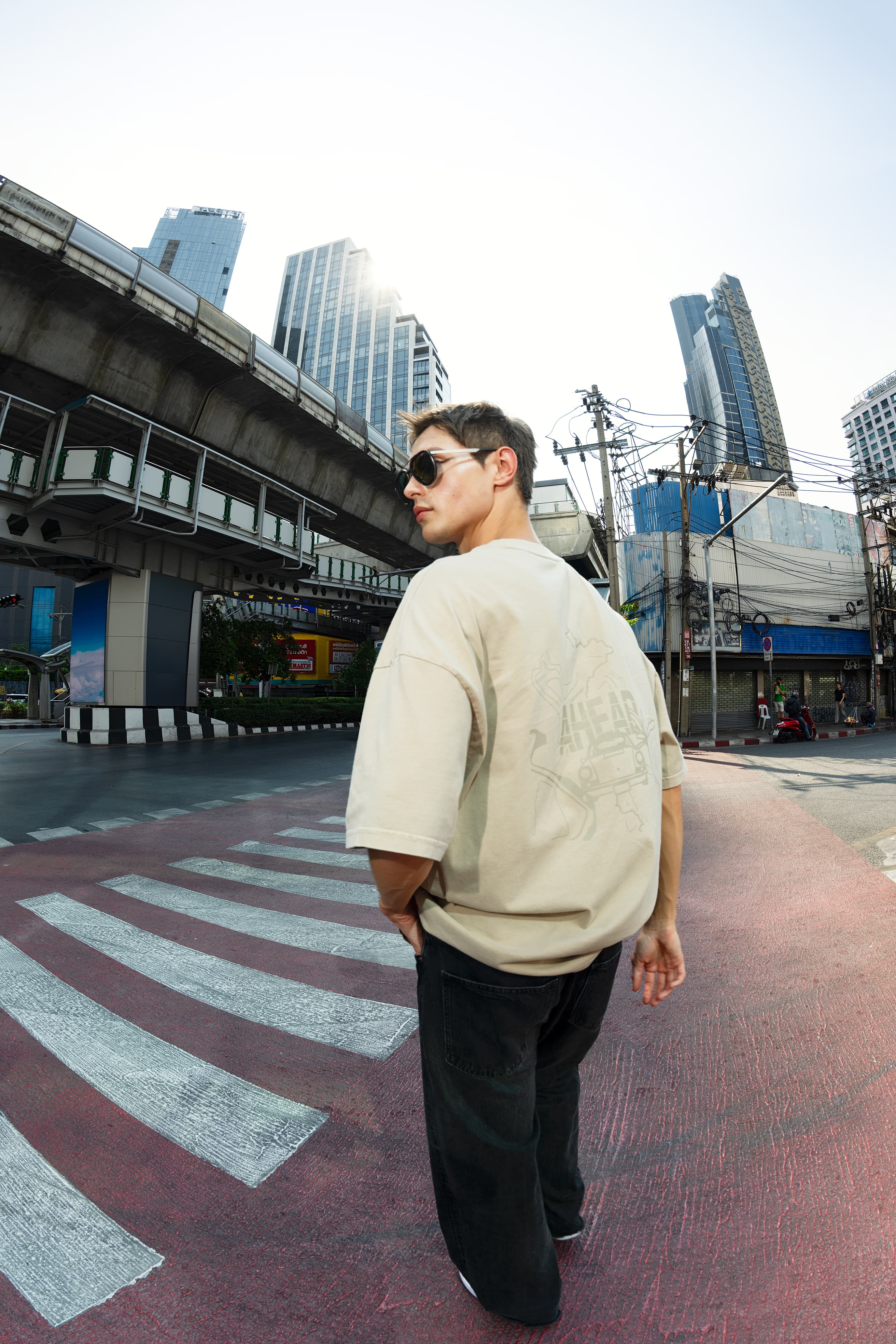 Uomo con occhiali da sole e T-shirt beige attraversa un passaggio pedonale; sullo sfondo grattacieli e linea sopraelevata.