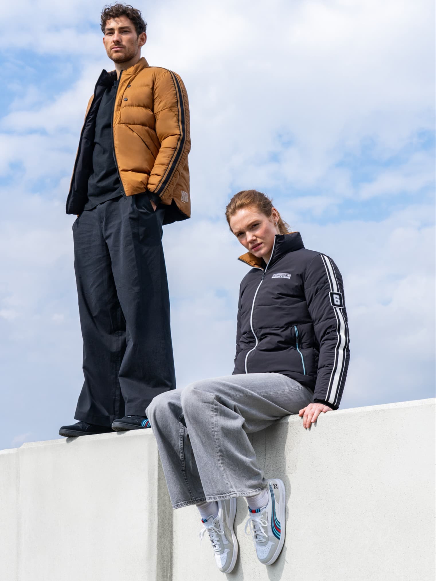 Twee mensen in casual jassen, één bruin en één zwart, staan en zitten op een witte muur tegen een bewolkte lucht.