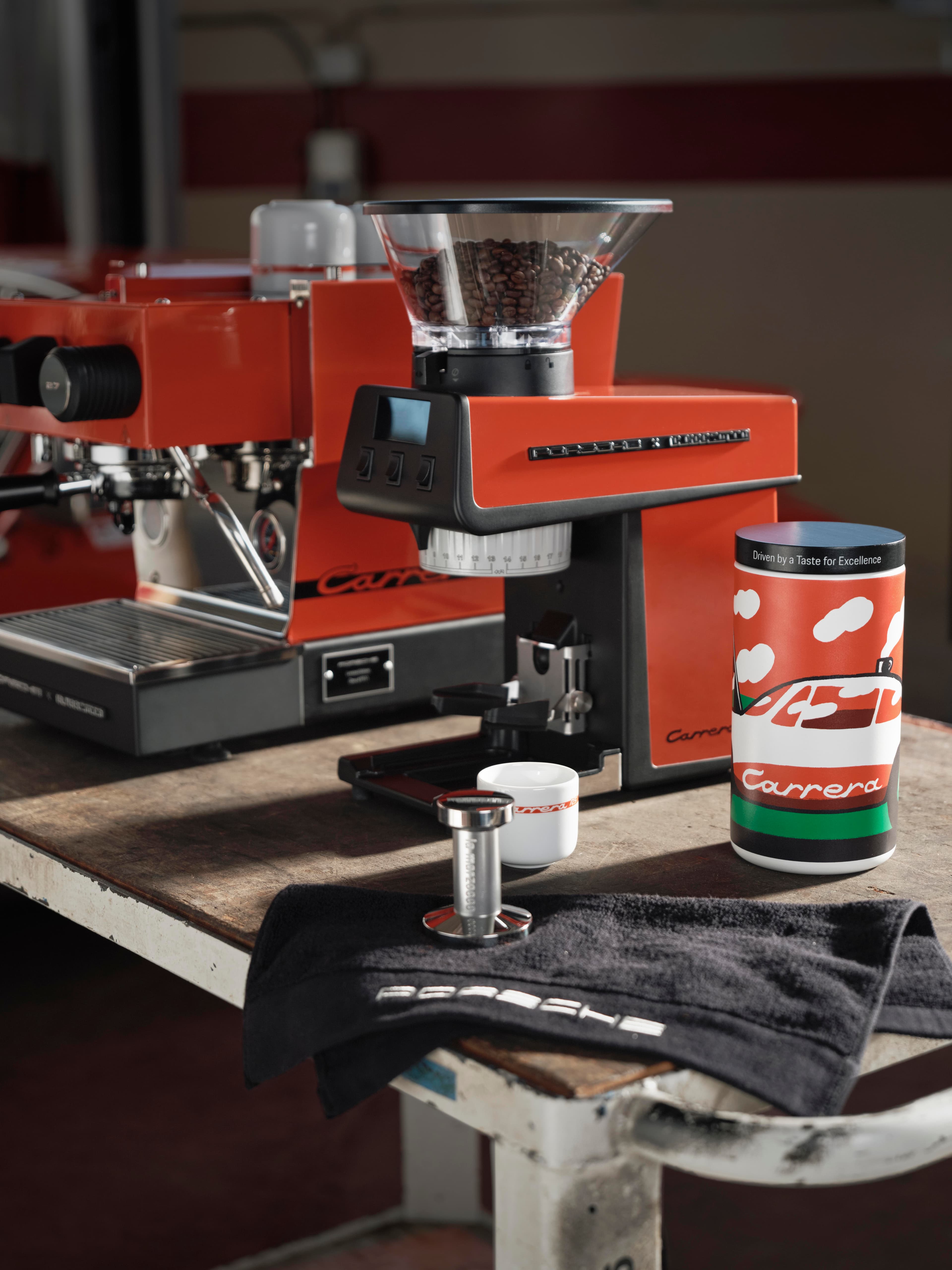 Ekspres do espresso i młynek do kawy z pojemnikiem na ziarna, obok puszki Carrera, tampera i czarnego ręcznika Porsche.