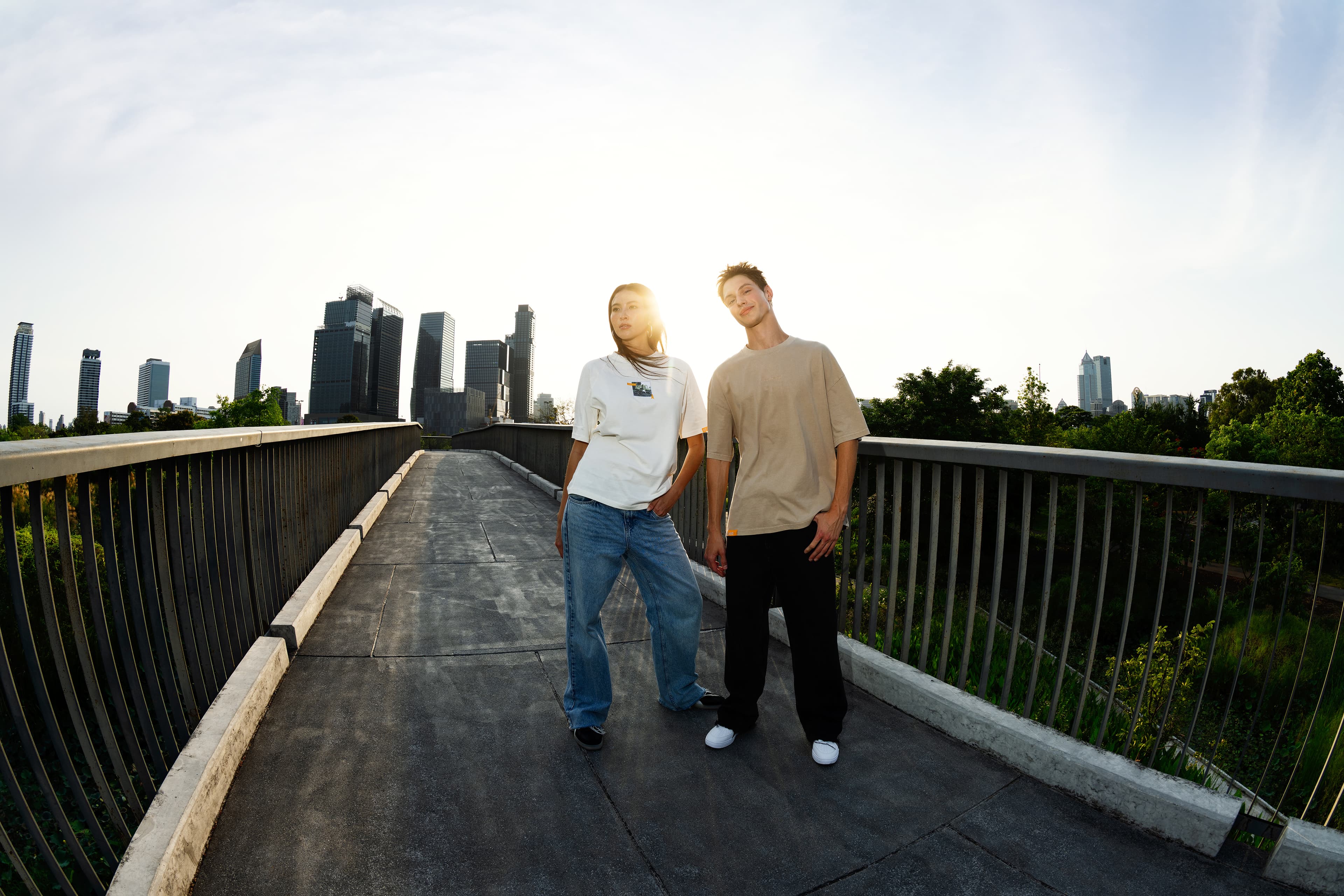 Two models on a bridge at sunset, wearing beige and white Porsche AHEAD T-shirts with jeans, city skyline in background.