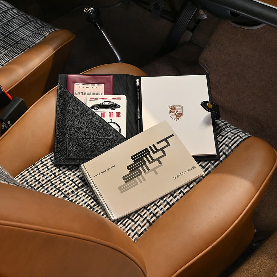 Porsche-Handbücher und Dokumente auf einem Hahnentritt-Autositz, mit einer schwarzen Lederhülle und einem weißen Notizbuch mit Logo.
