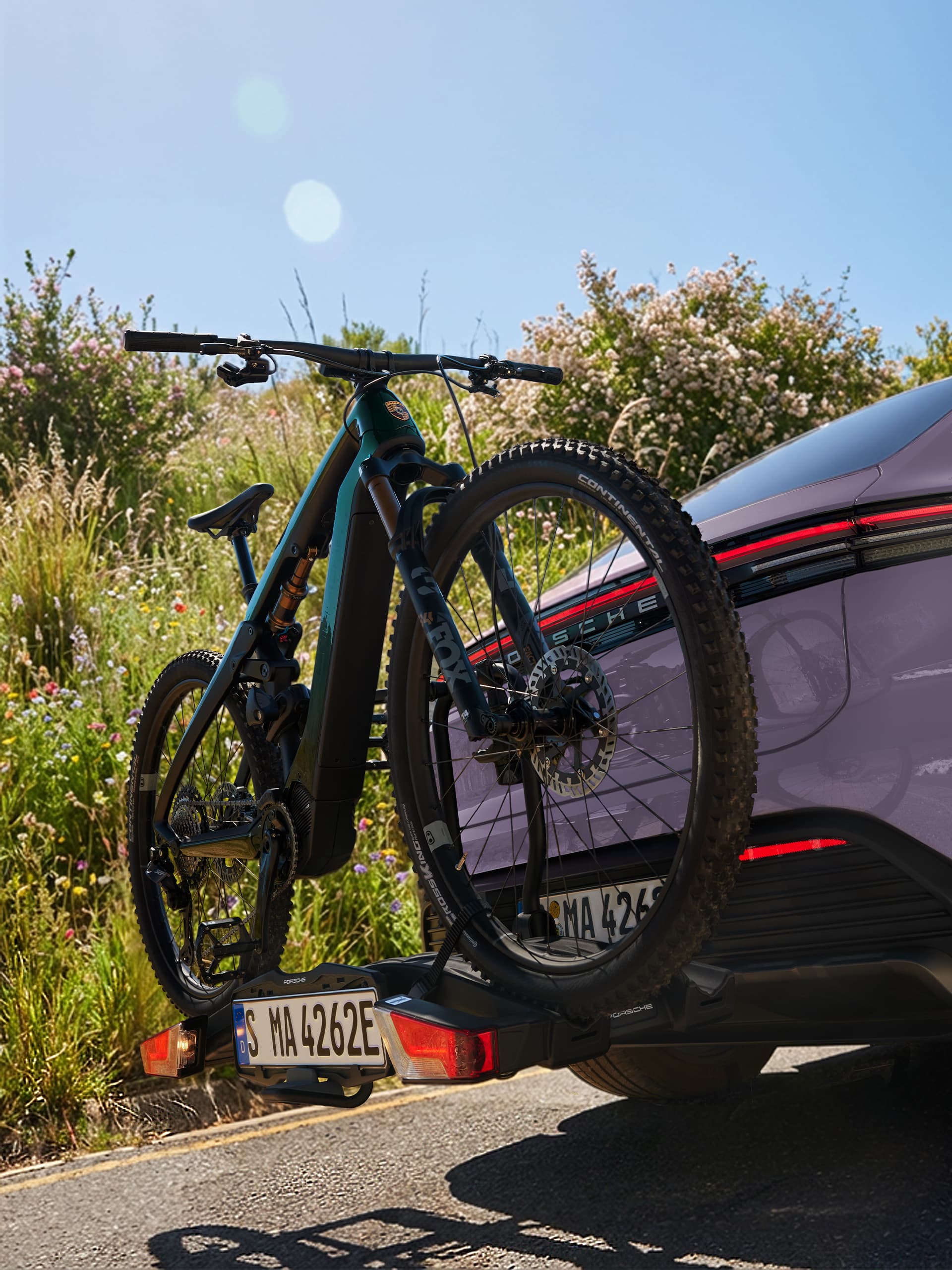 Fahrrad auf dem Heck eines lila Porsche montiert, vor einem sonnigen Hintergrund mit Grünflächen und Blumen.