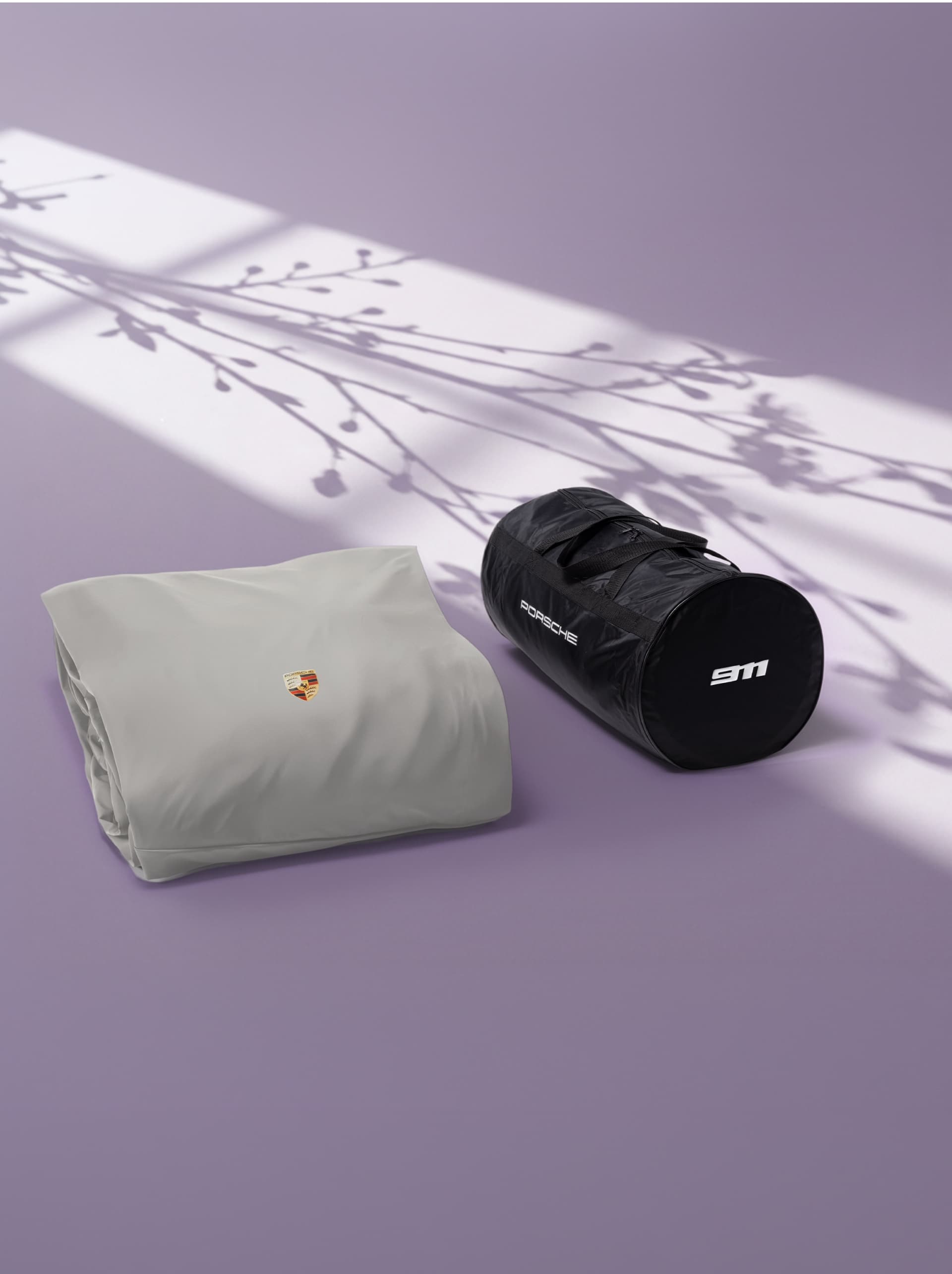 Housse Porsche grise et sac de sport noir sur une surface violette avec des ombres florales.