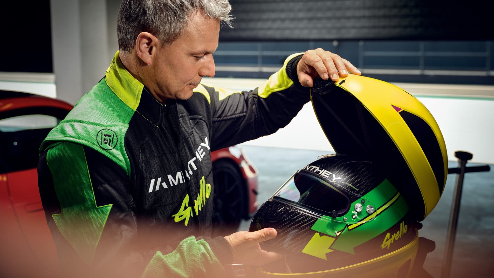 Hombre con traje de carreras sosteniendo un estuche de casco amarillo con un casco verde y negro adentro, coche rojo al fondo.
