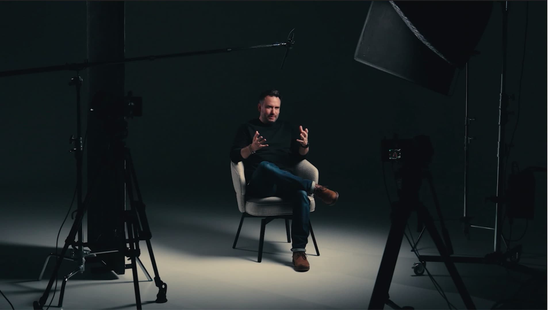 Mężczyzna w czarnym swetrze siedzi w fotelu w zaciemnionym studiu fotograficznym, otoczony sprzętem oświetleniowym i fotograficznym.