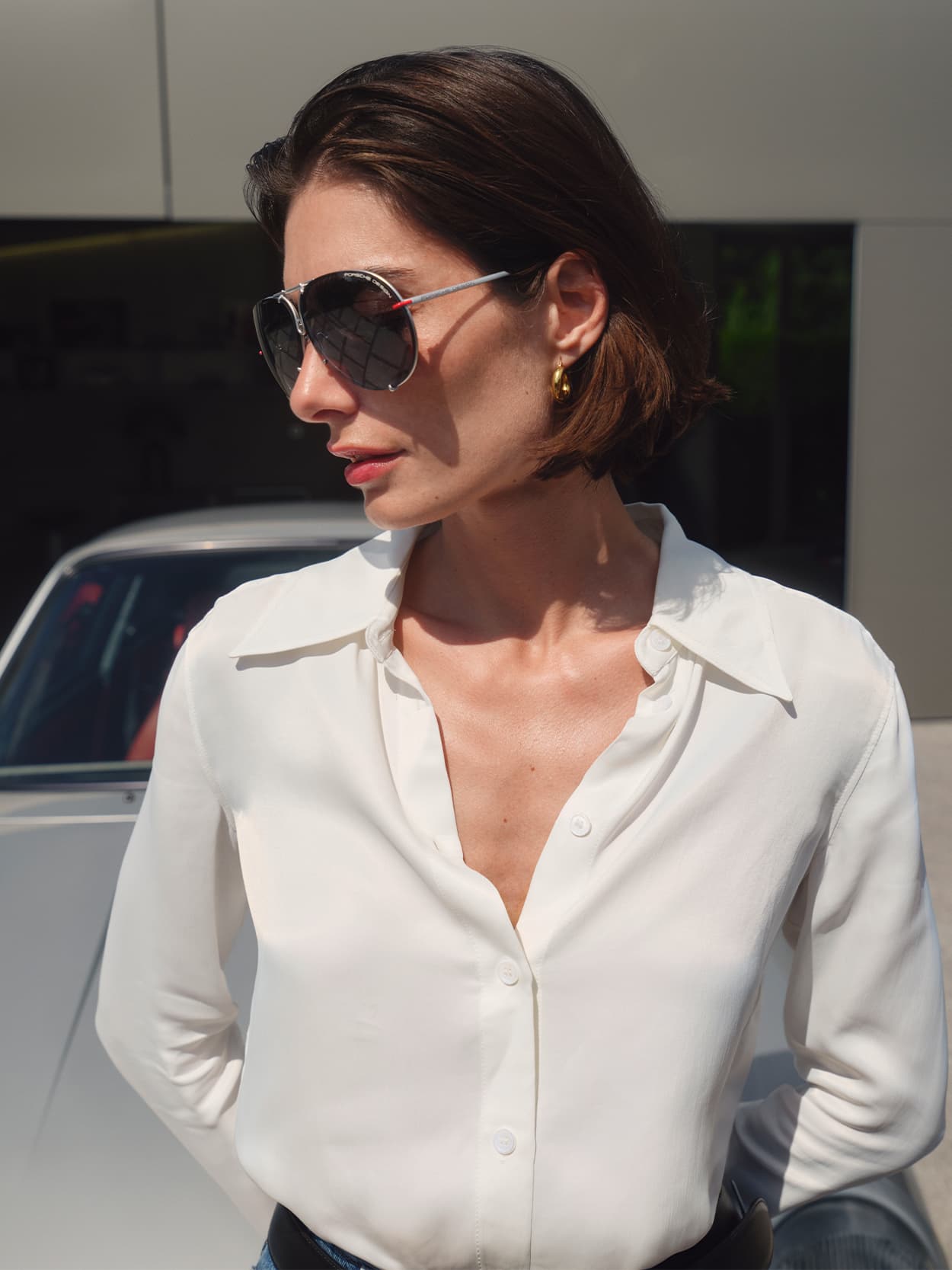A woman standing in front of a Porsche car, looking to her right and wearing Porsche Design Sunglasses.