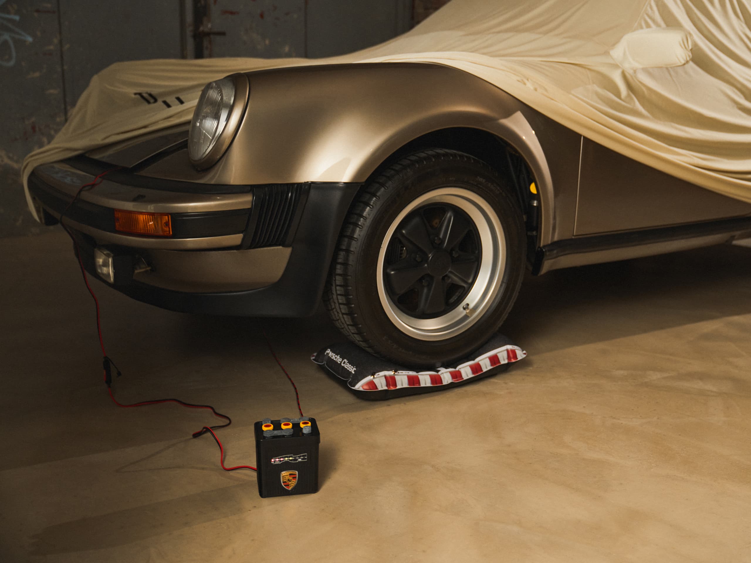 Klassischer Porsche, teilweise abgedeckt, goldfarben, mit angeschlossenem Batterieladegerät, auf Betonboden geparkt.