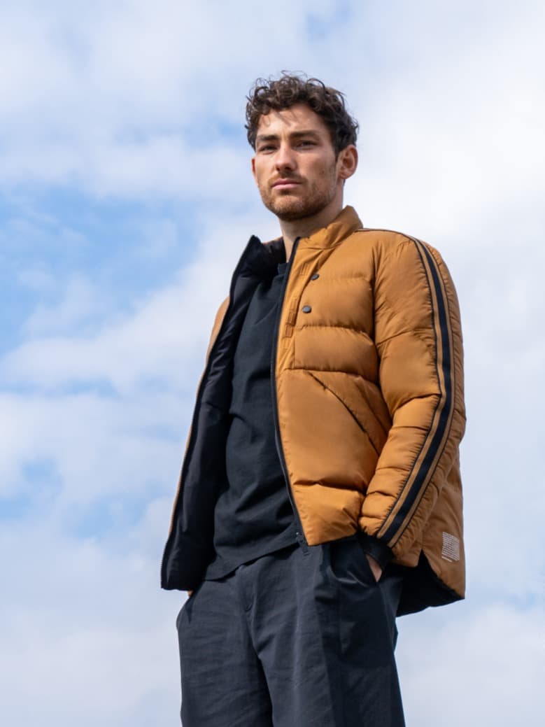 Man in a brown puffer jacket and black shirt stands against a cloudy sky, looking forward.