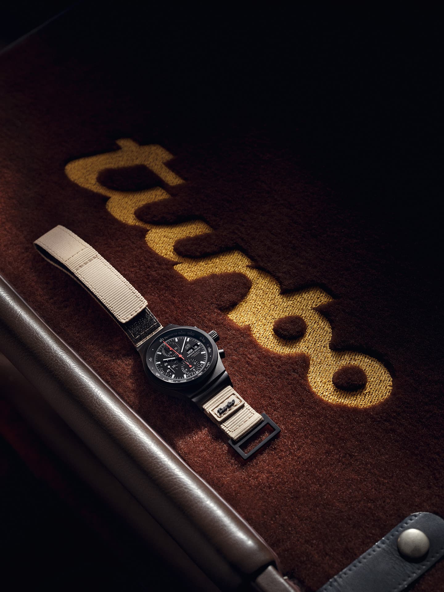 Czarny chronograf z beżowym tekstylnym paskiem na brązowej filcowej podkładce ze złotym haftem „turbo”.