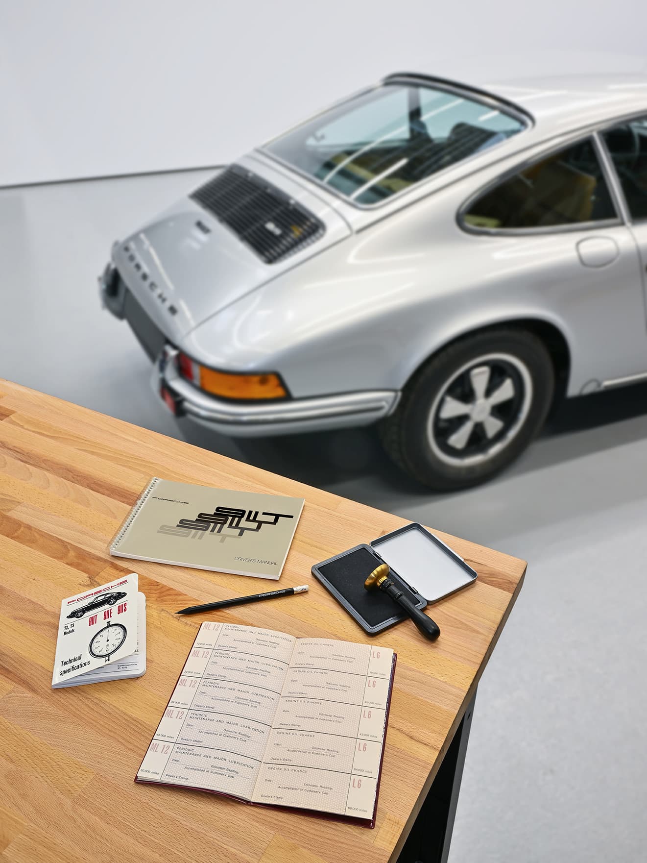 Wooden table with Porsche manuals, stamp, and service booklet; silver Porsche 911 in the studio background.
