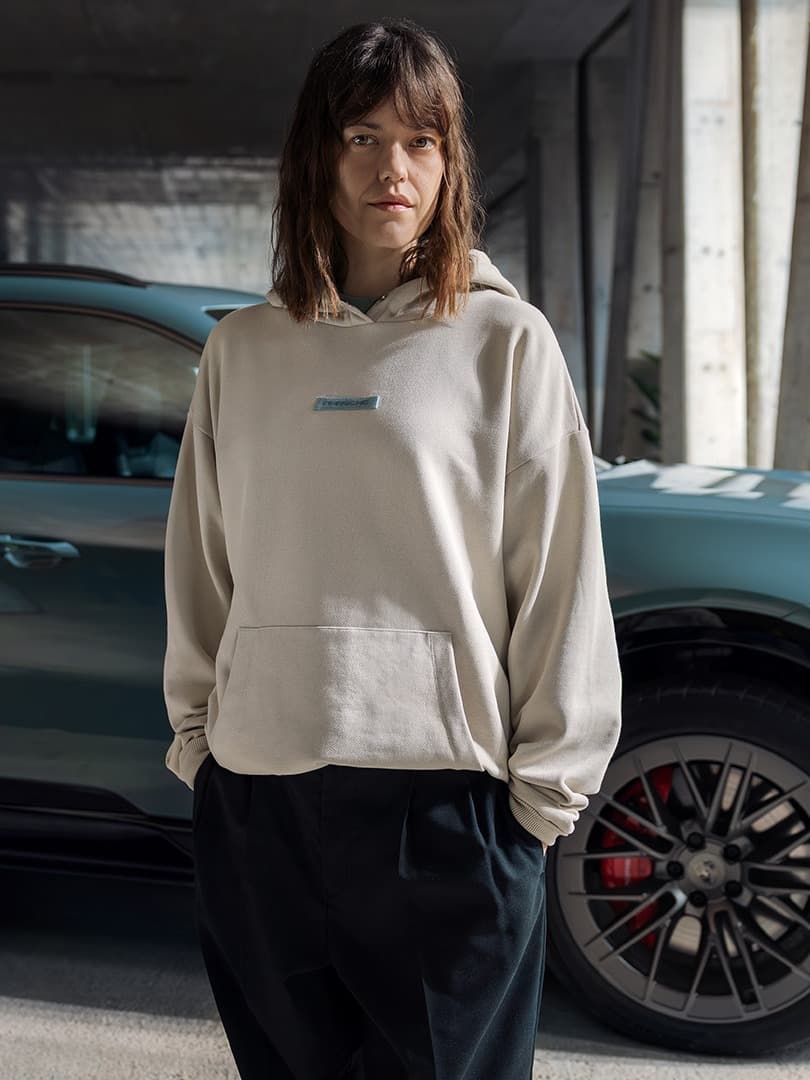 Woman in beige hoodie and man in blue shirt stand by a blue Porsche in a concrete garage, sunlight streaming in.