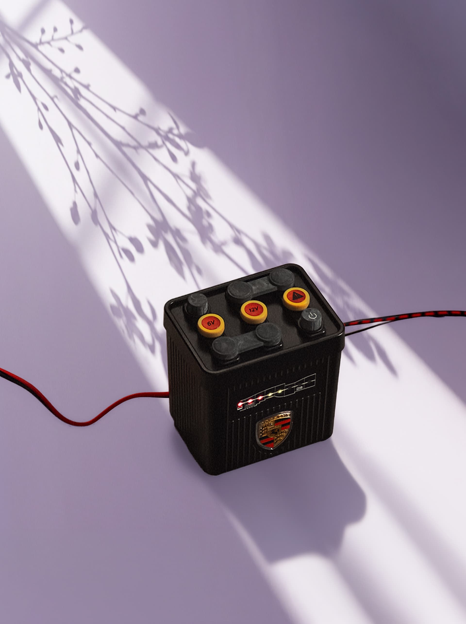 Black Porsche battery on a purple surface with shadowed branches, featuring red and yellow buttons and a logo.