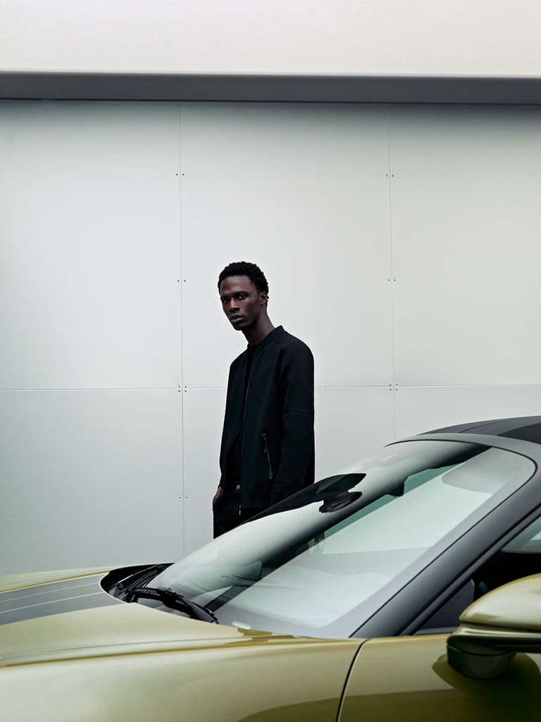 Hombre con chaqueta negra junto a un coche verde elegante frente a una pared blanca minimalista.