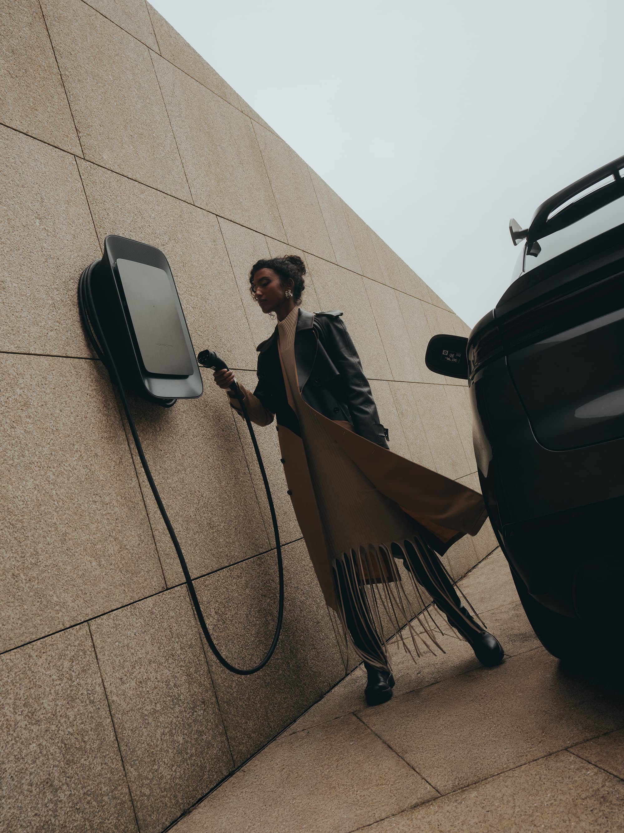 Person charging electric car at wall-mounted station, wearing long coat, next to a modern building.