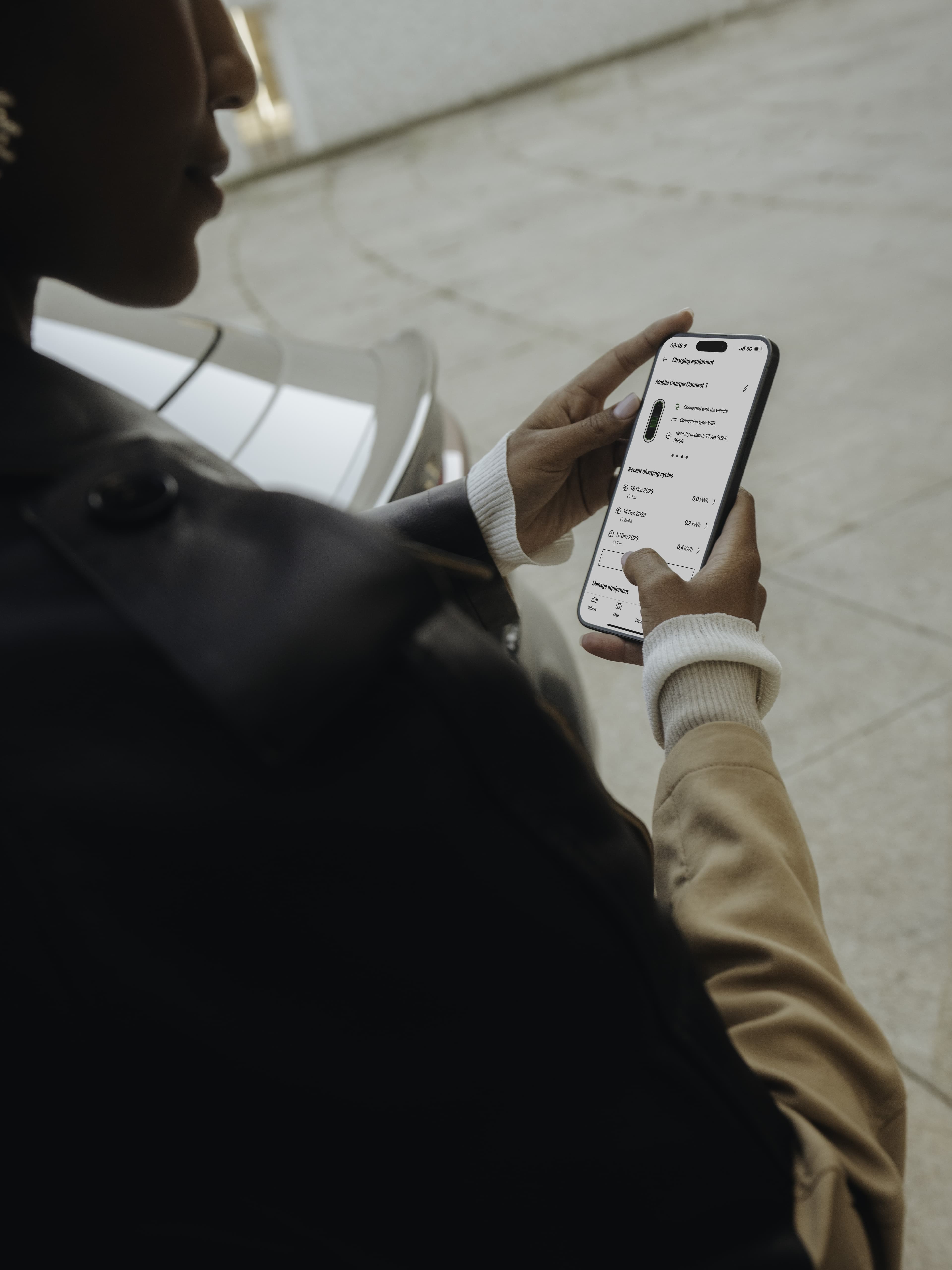 Person in a jacket using a smartphone app near a parked car, displaying charging details on the screen.