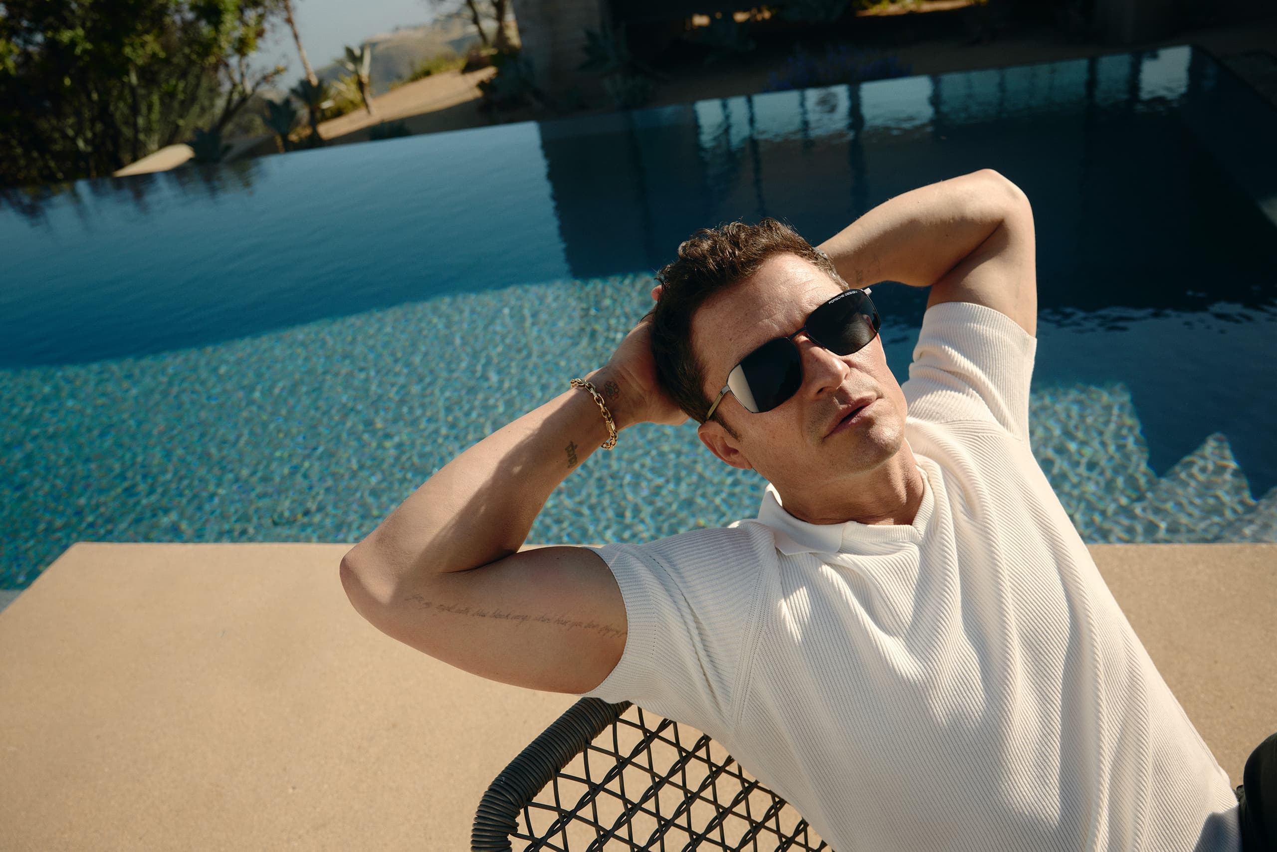 Orlando Bloom wears the Porsche Design Motion Eyewear series, sits on a chair, leans back, and looks up. A pool can be seen in the background.